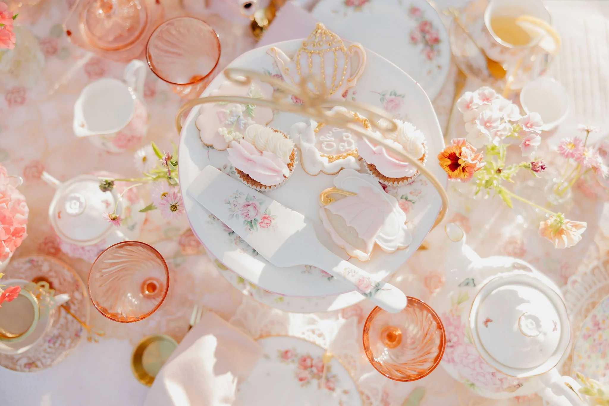 Elegant dessert table with ornate cookies, pink and gold decorations, and floral accents on fine china.