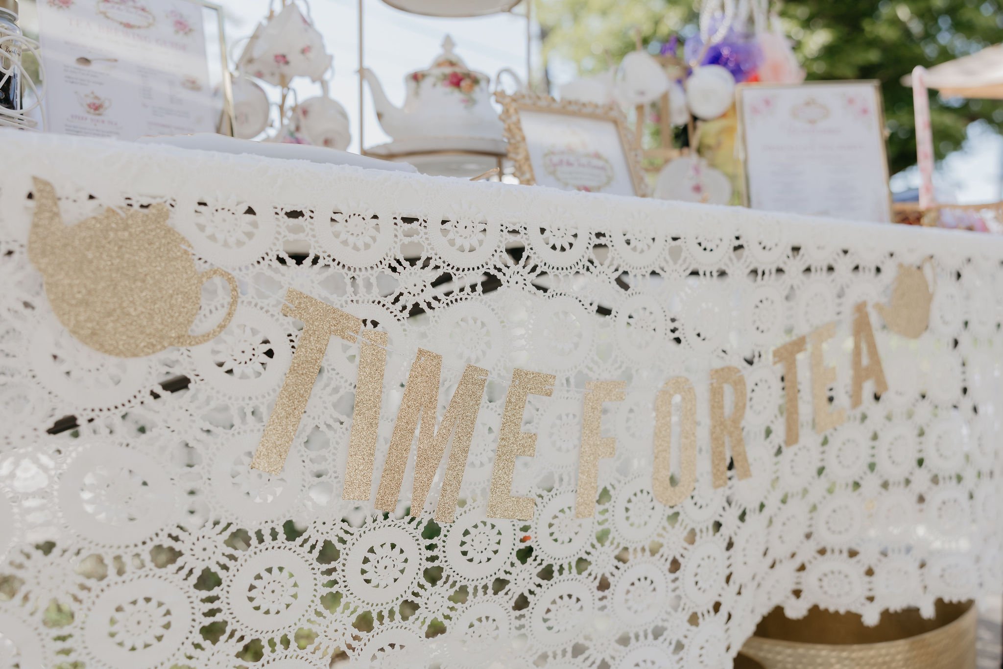 Close up of a white lace tablecloth with gold glittery cutout signs hanging on it, at an outdoor tea party or celebration with vintage teapots and floral decorations in the background.