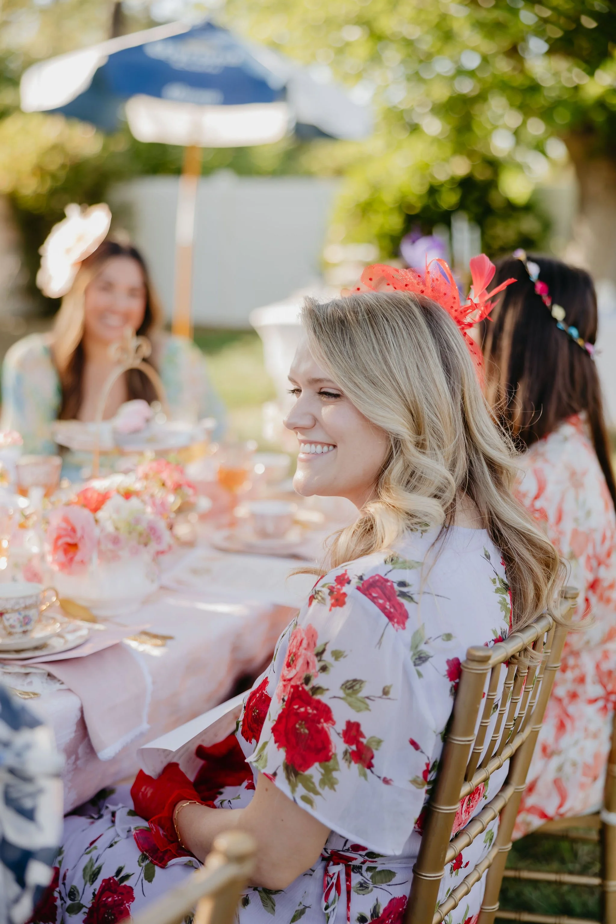 Smiling blonde woman with floral dress and red headband sitting at outdoor tea party table with other women, colorful flowers, teacups, and a tablecloth, in a garden with green trees.