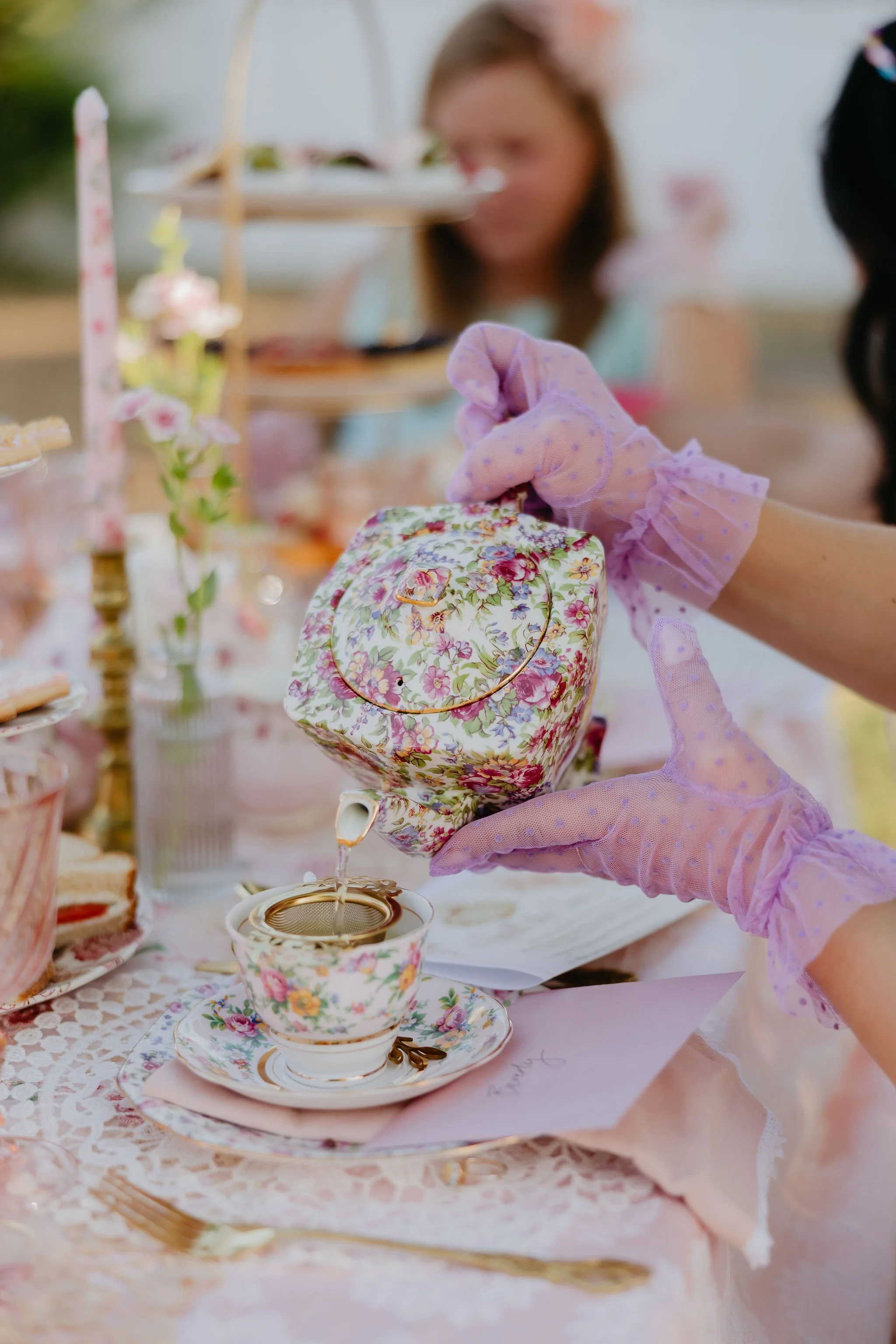 A person wearing pink gloves is pouring tea into a floral china cup at a tea party. The table is decorated with flowers, teacups, and desserts, with a blurred girl in the background.