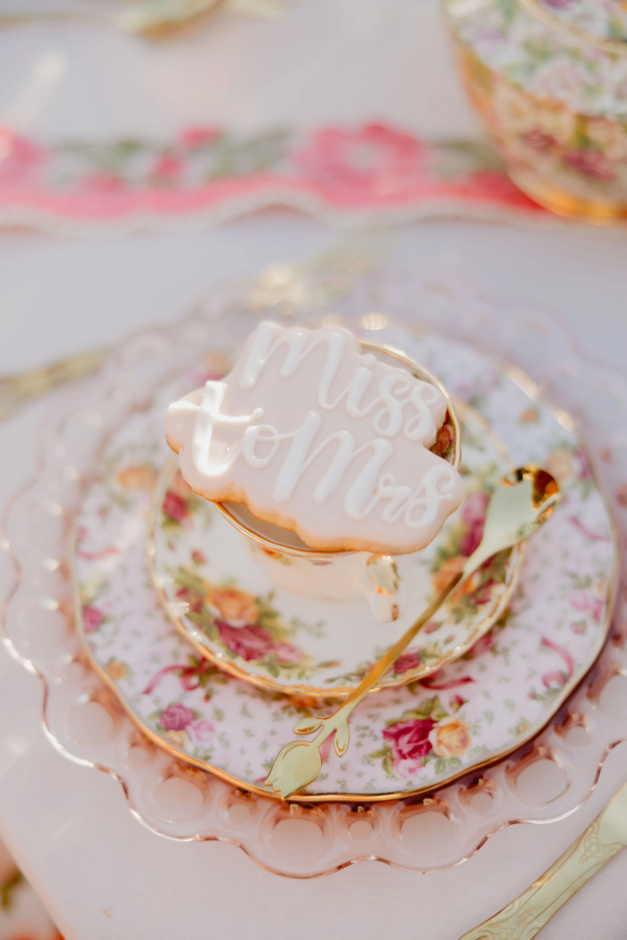 A floral patterned teacup with a matching saucer, topped with a heart-shaped cookie that has white icing and the words "Miss You" written in white icing.