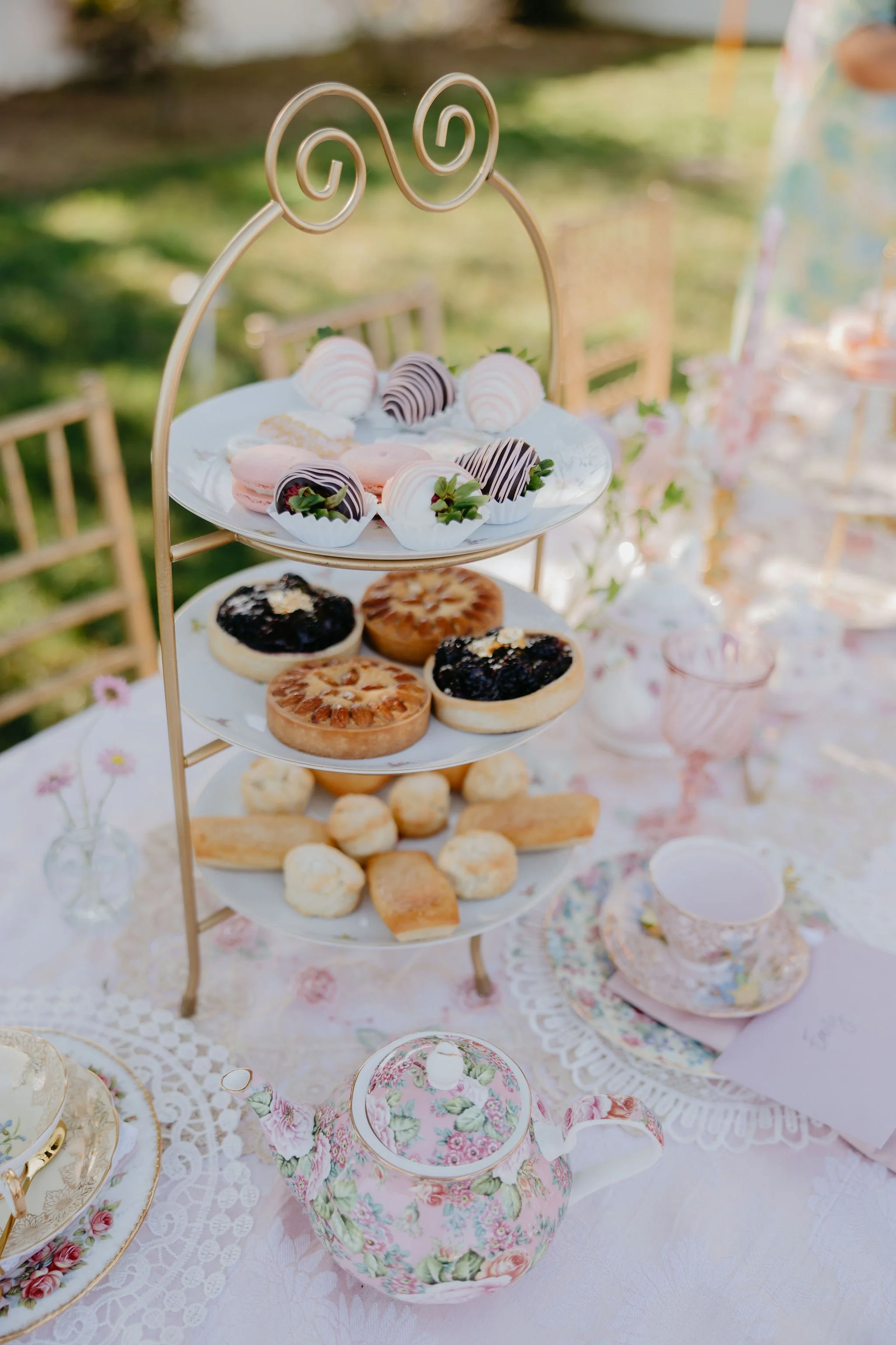 A three-tiered stand with assorted pink and chocolate dipped desserts, fruit tarts, and small cookies on a floral and lace tablecloth, outdoors with blurred greenery in the background.
