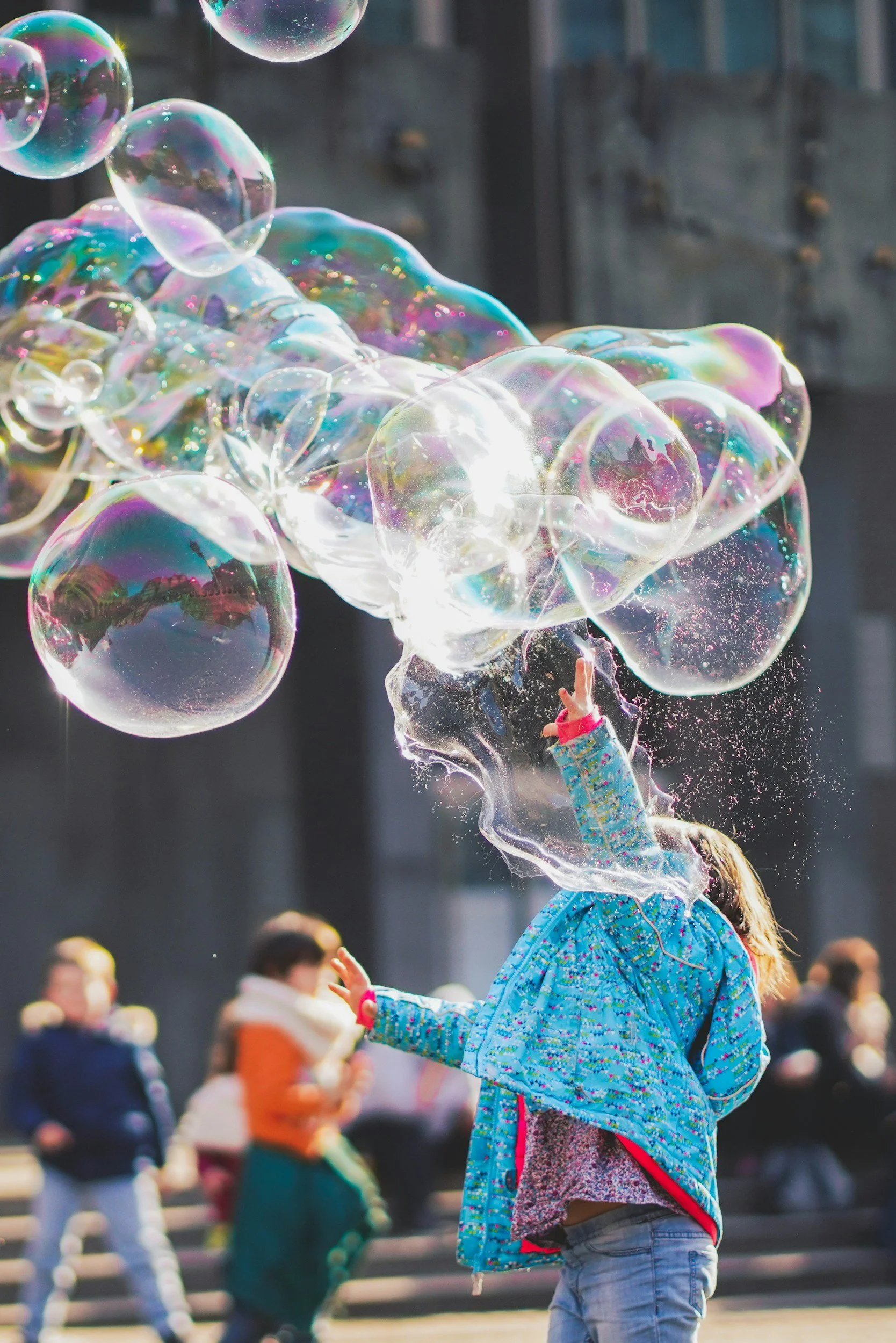 Child in a blue jacket with colorful patterns reaching out to soap bubbles floating in the air during daytime in a crowded outdoor area.