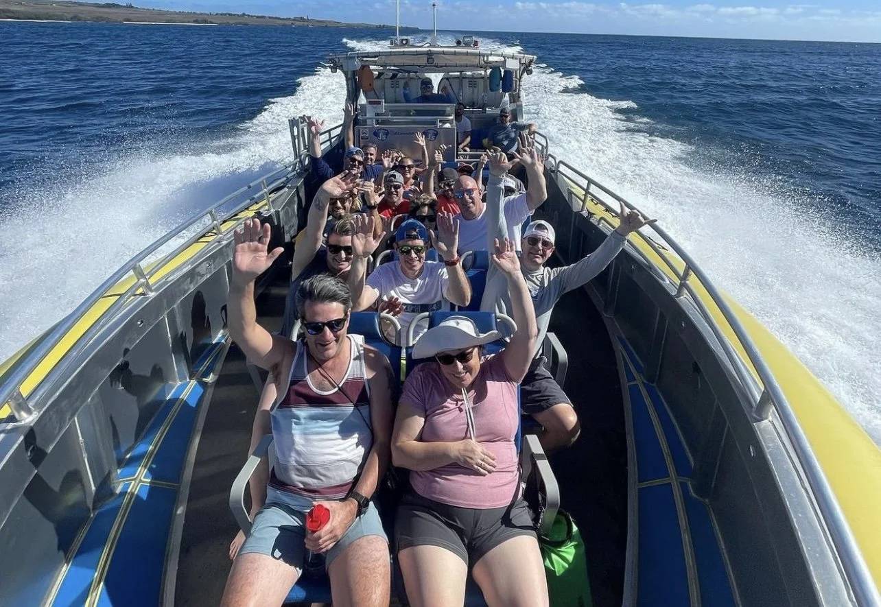A group of people enjoying a boat ride on the water, waving at the camera with blue ocean and sky in the background.