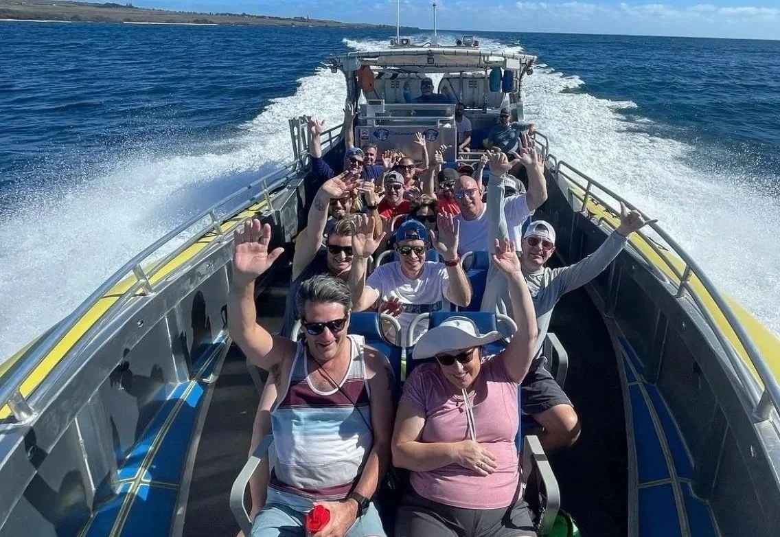 Group of people on a boat enjoying sunny weather, some waving at the camera, with water and shoreline visible in the background.