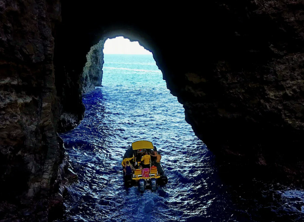 A boat moving through a sea cave with a rocky opening and sunlight shining through, revealing blue water.