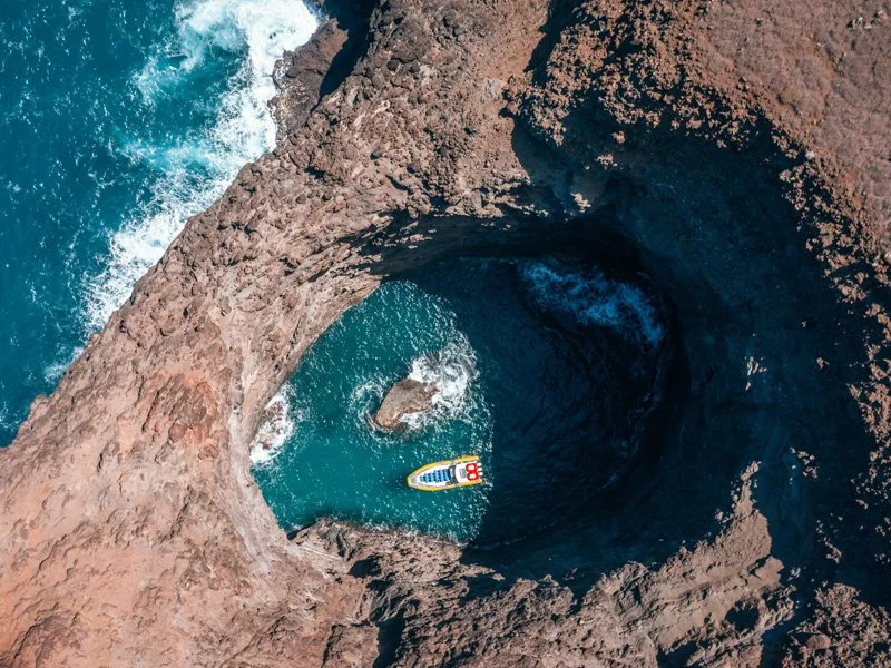 Aerial view of a large sea cave with a colorful paddleboarder inside, surrounded by rocky cliffs and deep blue ocean water.