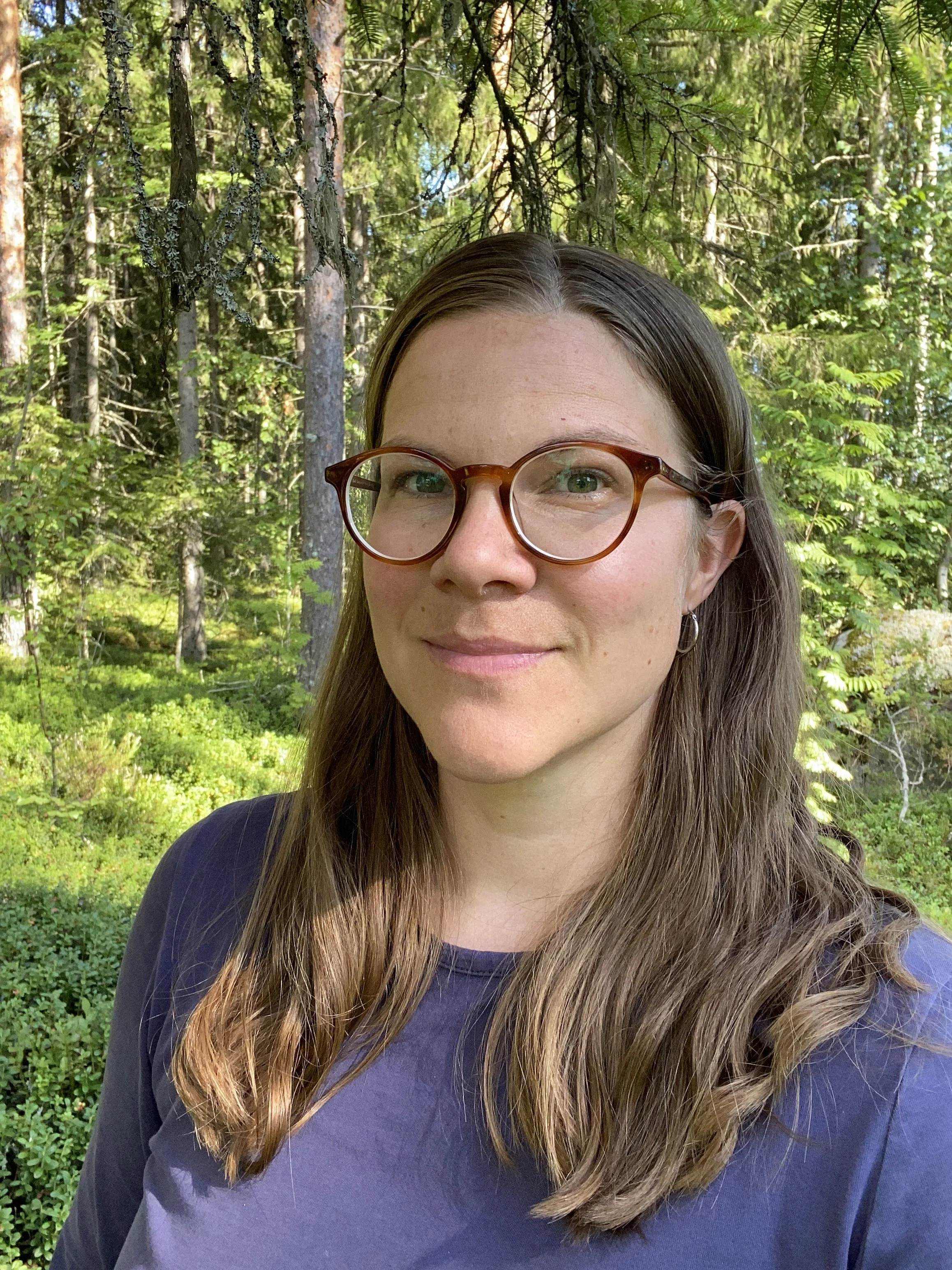 A woman with long, wavy light brown hair and glasses with round frames, standing outdoors in a forest with green trees and foliage in the background.