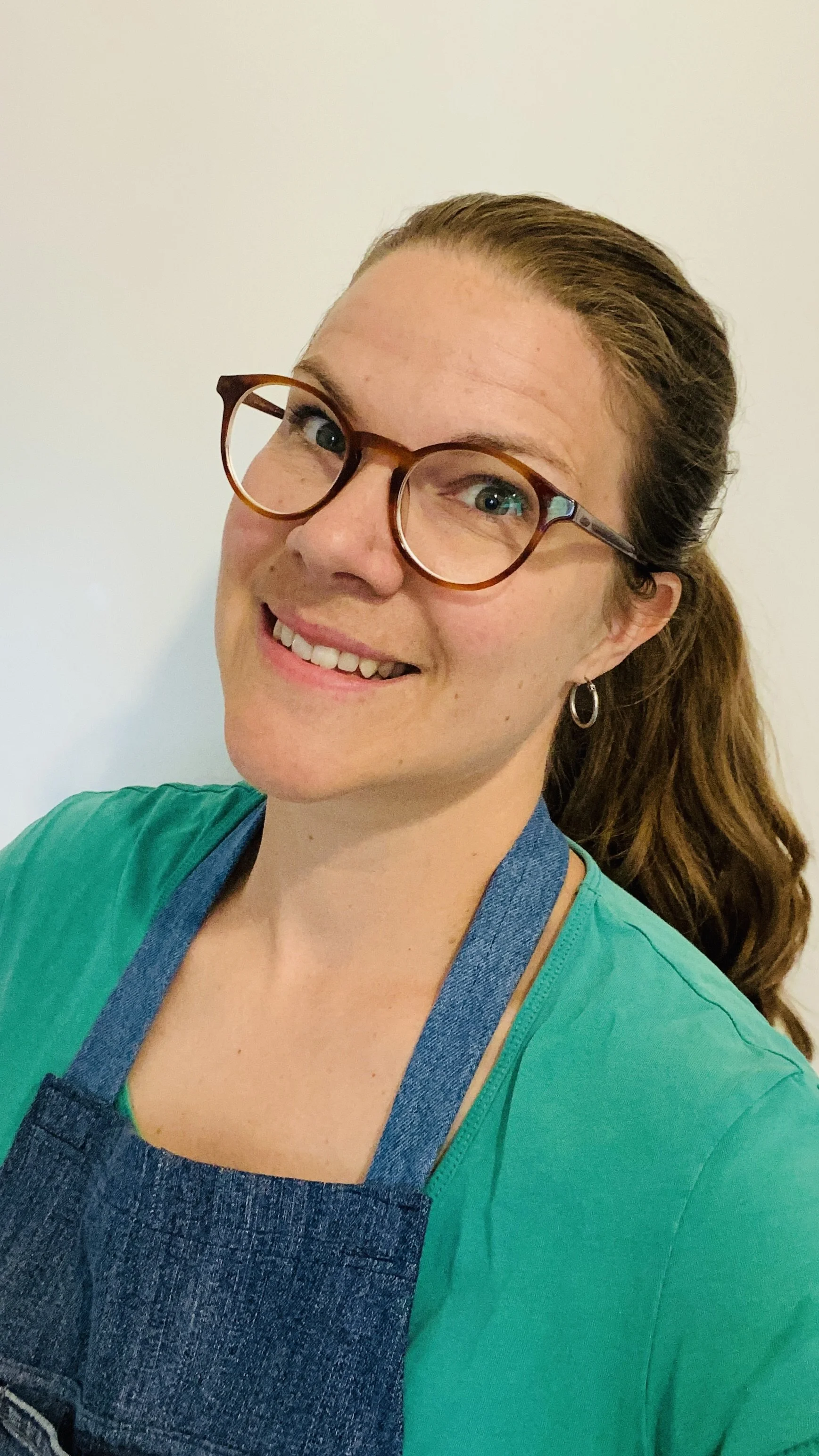 A woman with brown hair tied back, wearing glasses, earrings, a green shirt, and a denim apron, smiling with a plain wall in the background.