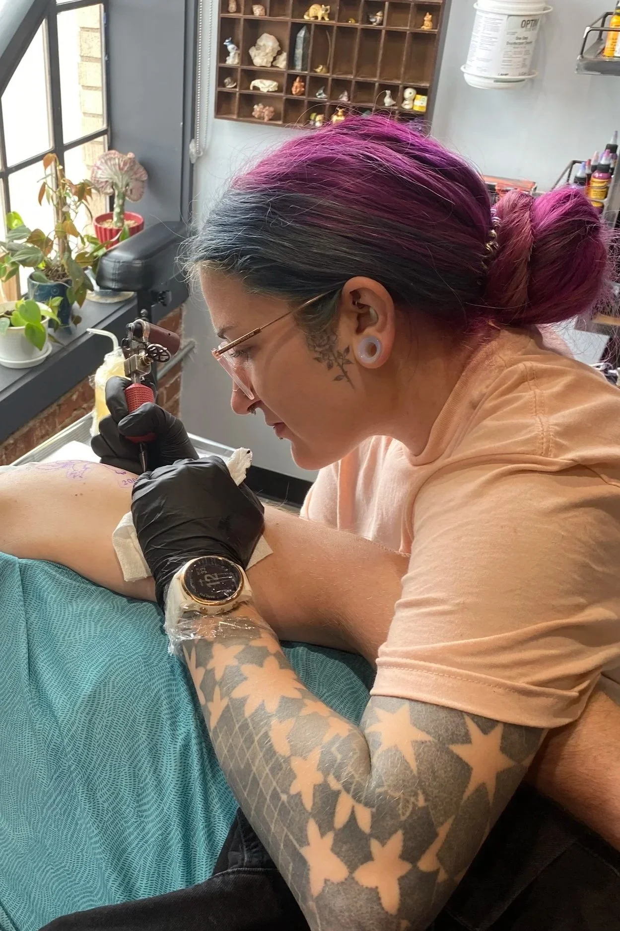 A tattoo artist with purple pink hair, tattoos on her arms and neck, wearing black gloves, is giving a tattoo to a client in a tattoo studio. The studio has plants near a window, a shelving unit with art supplies, and a wall with small figurines or sculptures.
