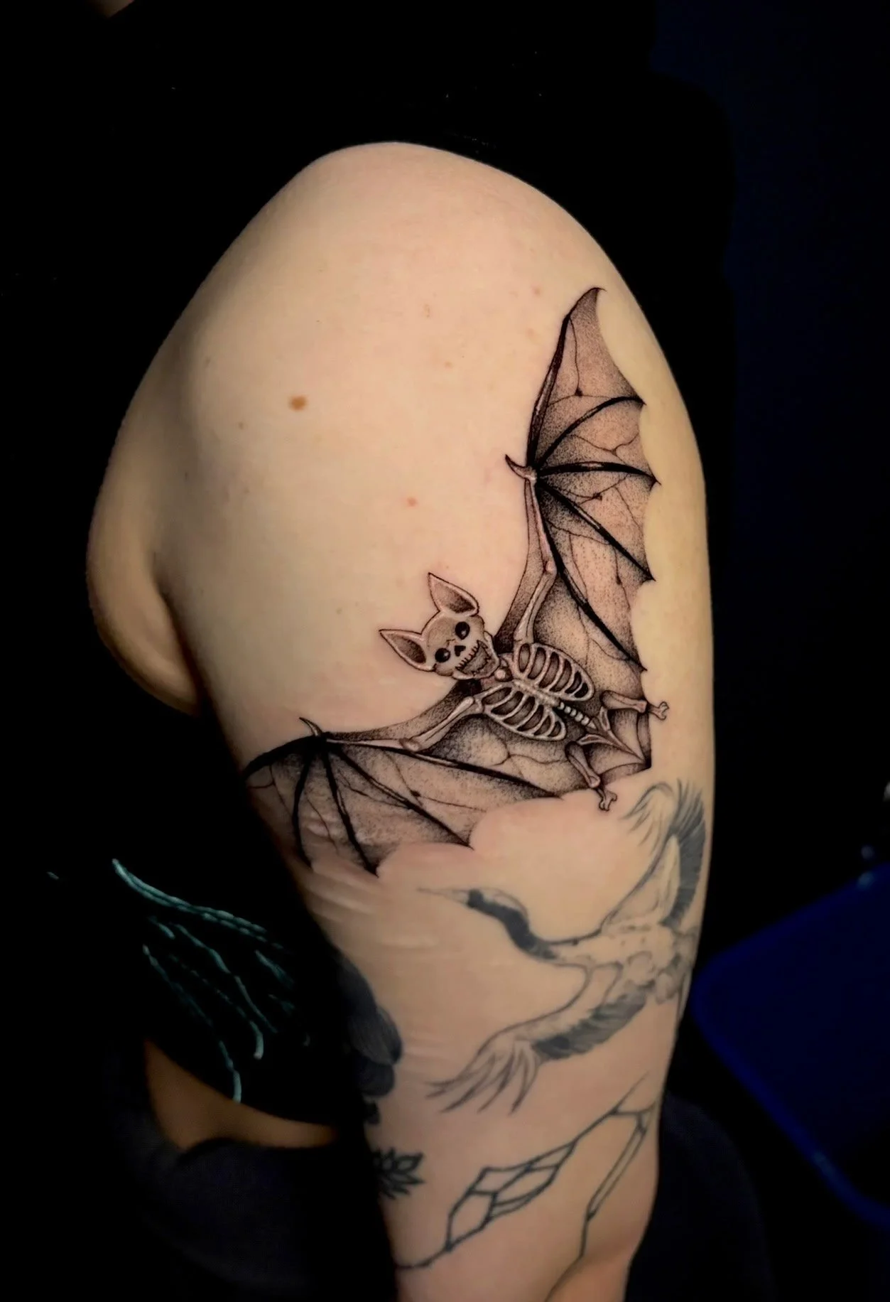 Tattoo of a cartoon-style bat skeleton with wings spread, on a person's upper arm.