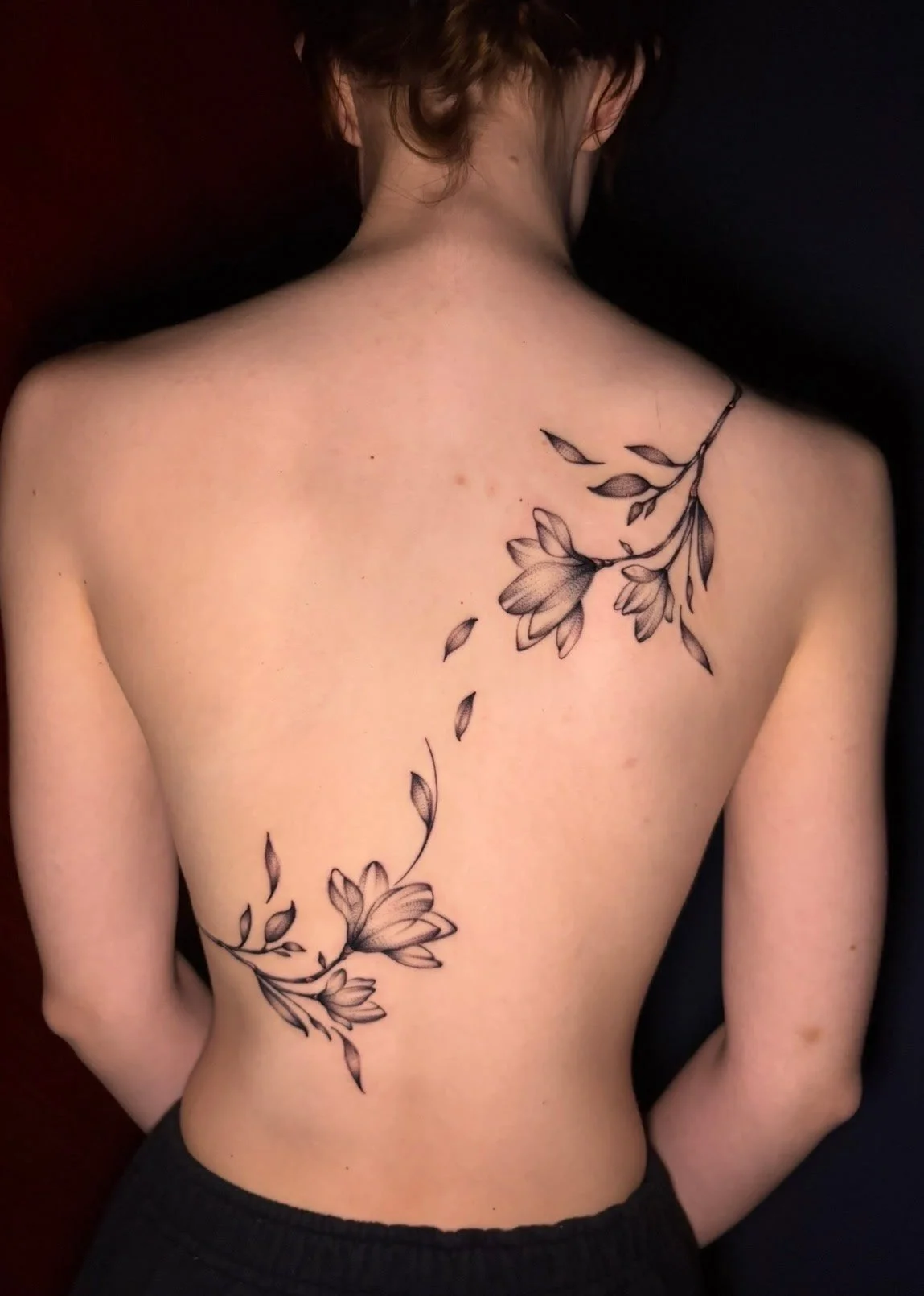 A woman with a floral tattoo on her back and shoulder, featuring black ink flowers and leaves with some petals falling.