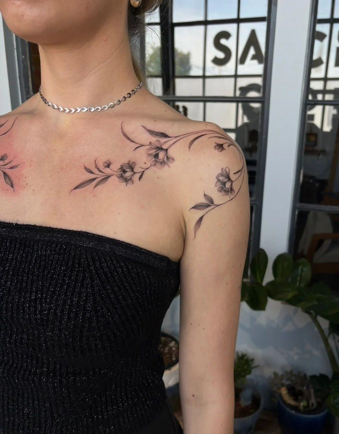 A woman showing her shoulder tattoo of a floral branch with black and gray flowers and leaves.