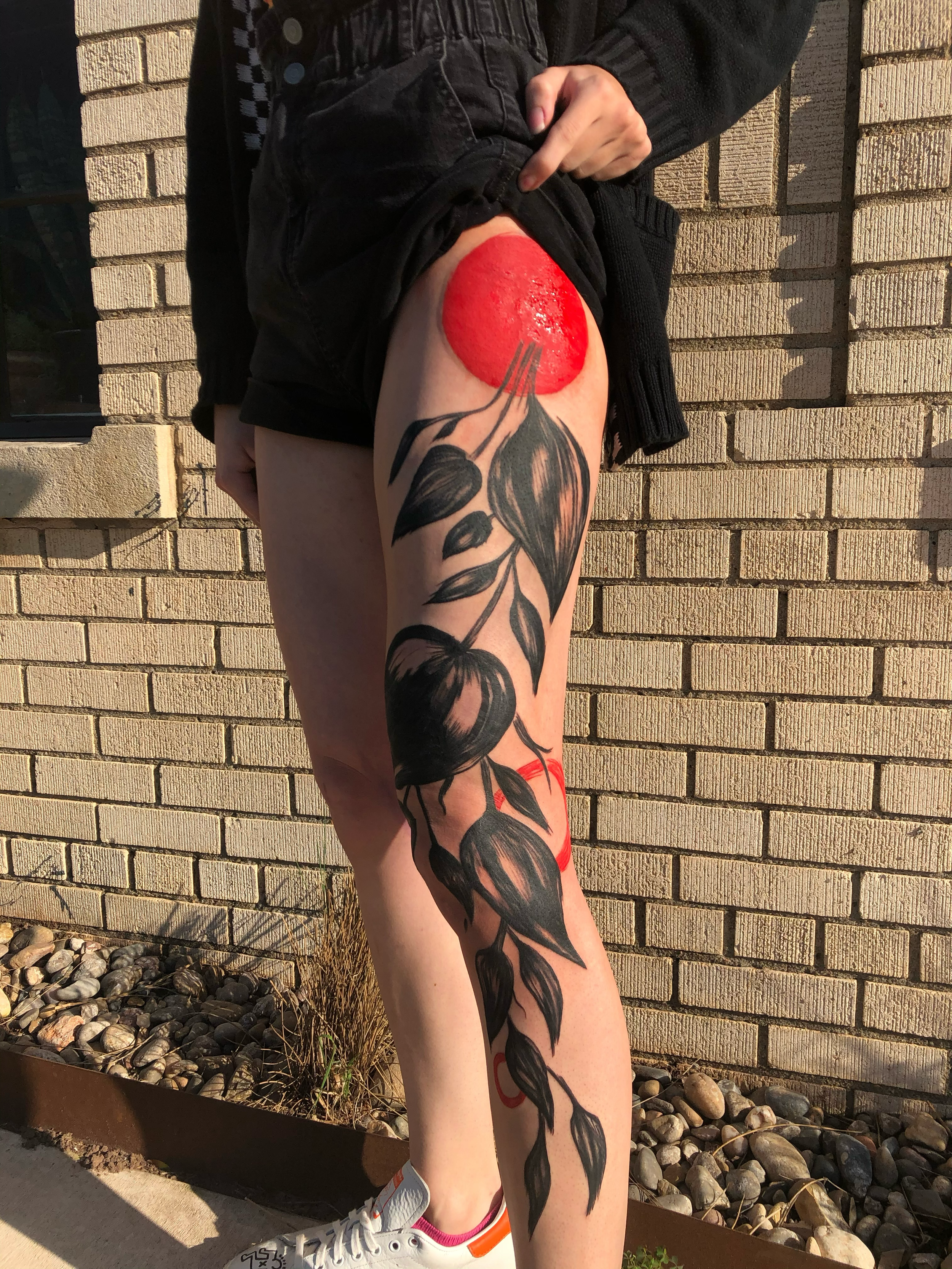 A person standing outdoors against a brick wall, showing their thigh with a large tattoo of a black and gray botanical design. They are lifting their black shorts to reveal a red circular paint or makeup mark on the upper thigh.