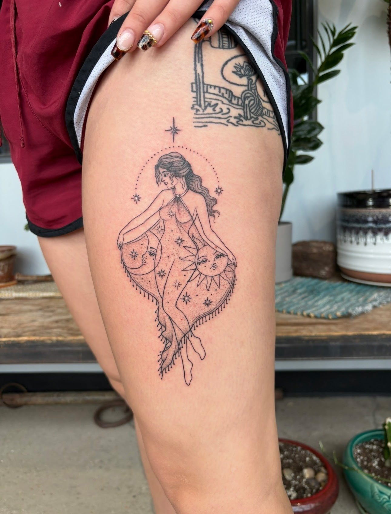 Tattoo of a woman with long wavy hair, surrounded by stars, with celestial faces of the moon and sun, on a person's thigh.