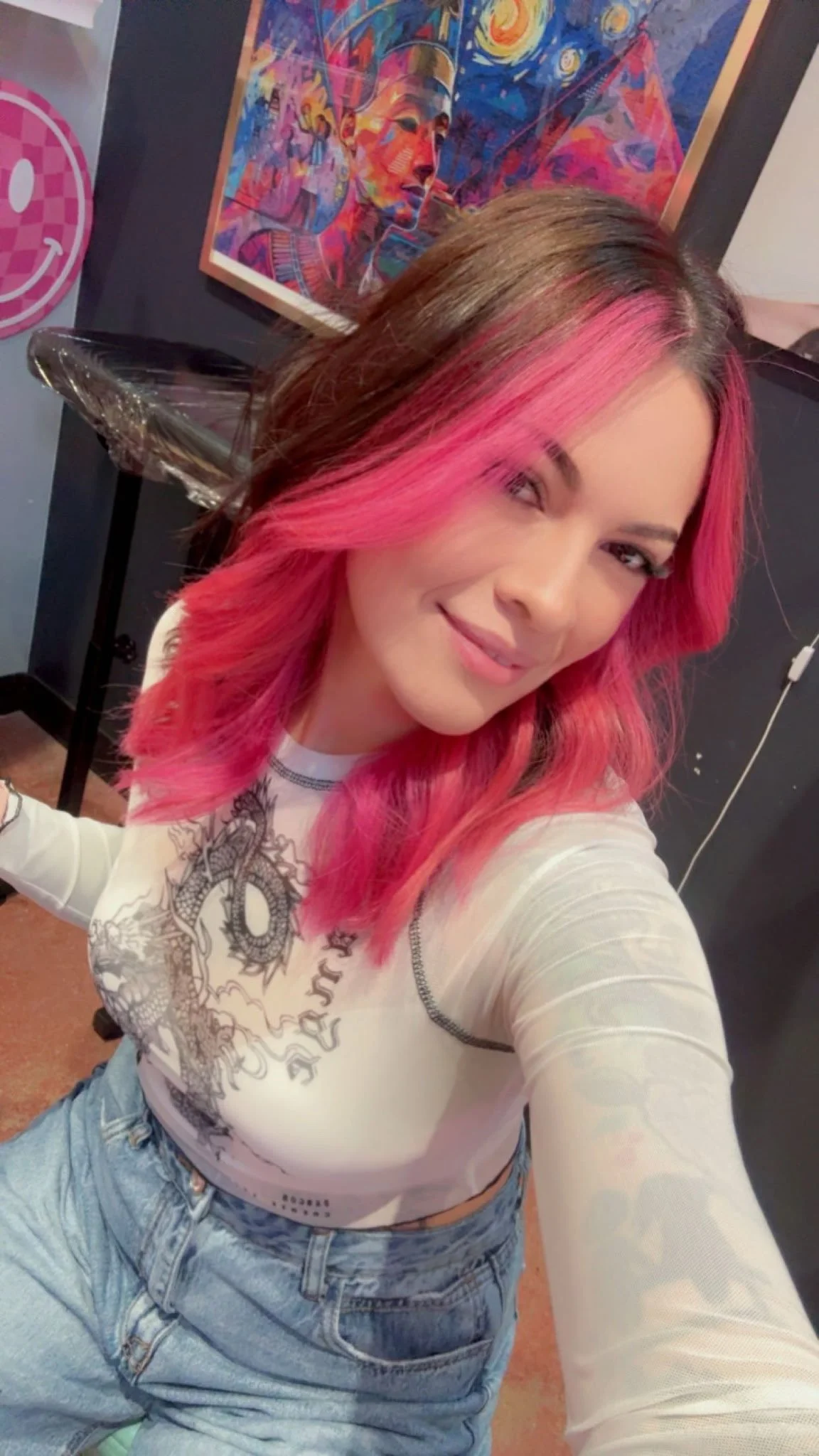 A woman with pink and brown hair taking a selfie in a room with a colorful abstract painting and a pink smiley face decoration on the wall behind her.