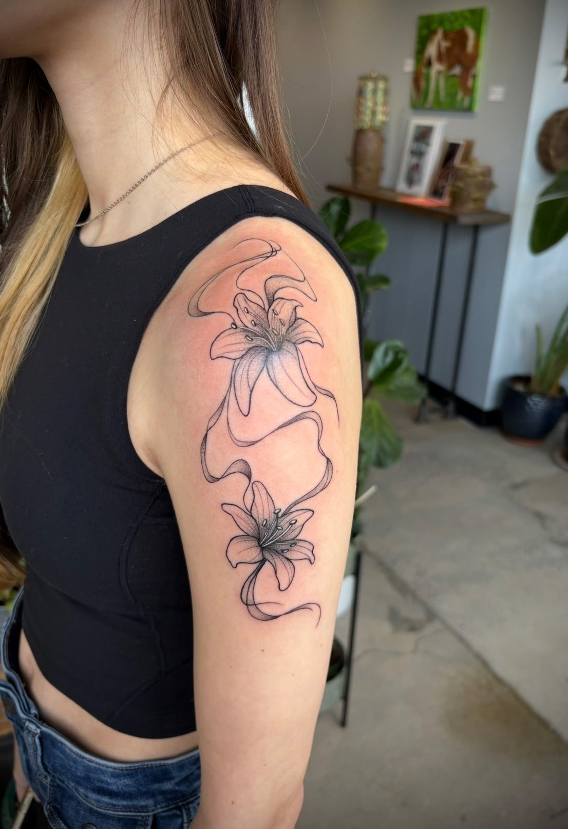 A woman with a black tank top and jeans showing a floral tattoo on her upper arm. The tattoo features two large lily flowers connected by flowing lines. The background includes indoor plants, artwork, and a table with books and decor.