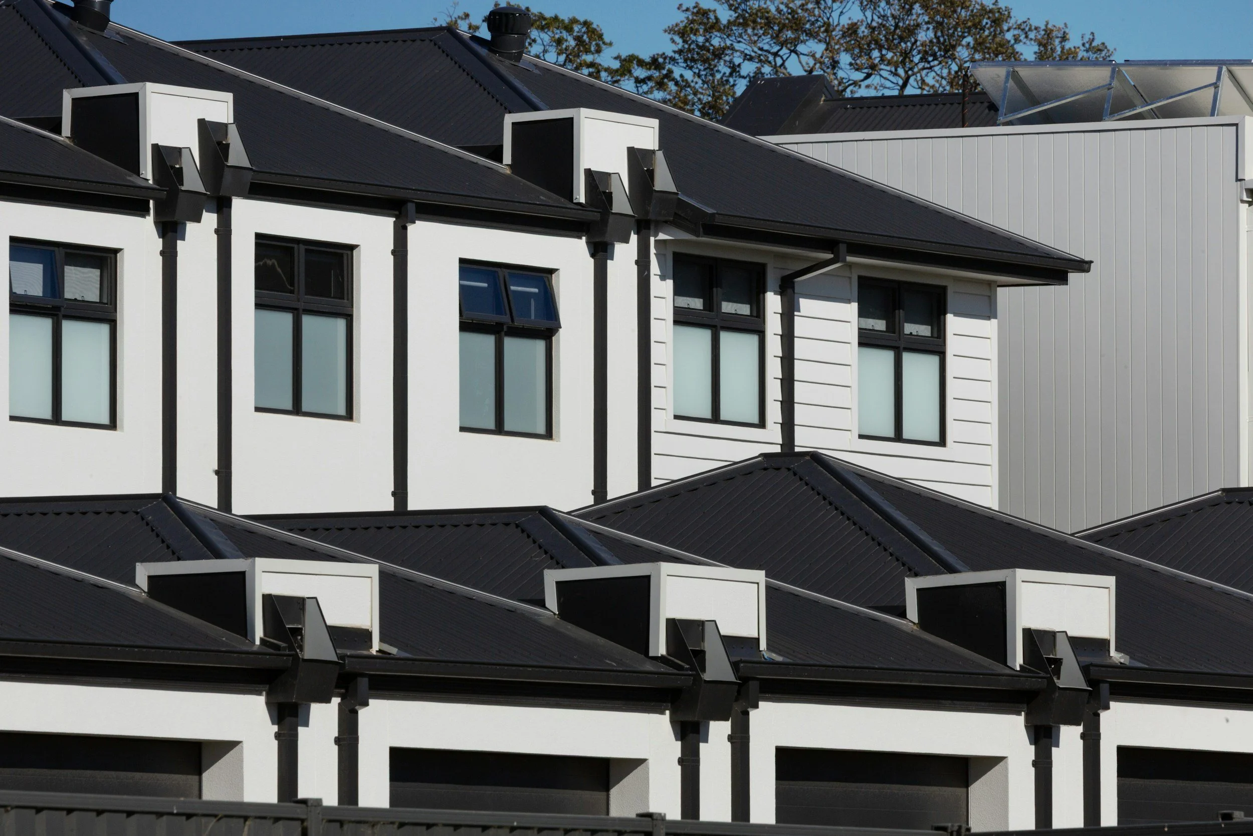 Close-up view of the rooftops of modern senior living or memory care buildings with black metal roofs, white walls, and black-framed windows under a clear blue sky with some trees in the background.