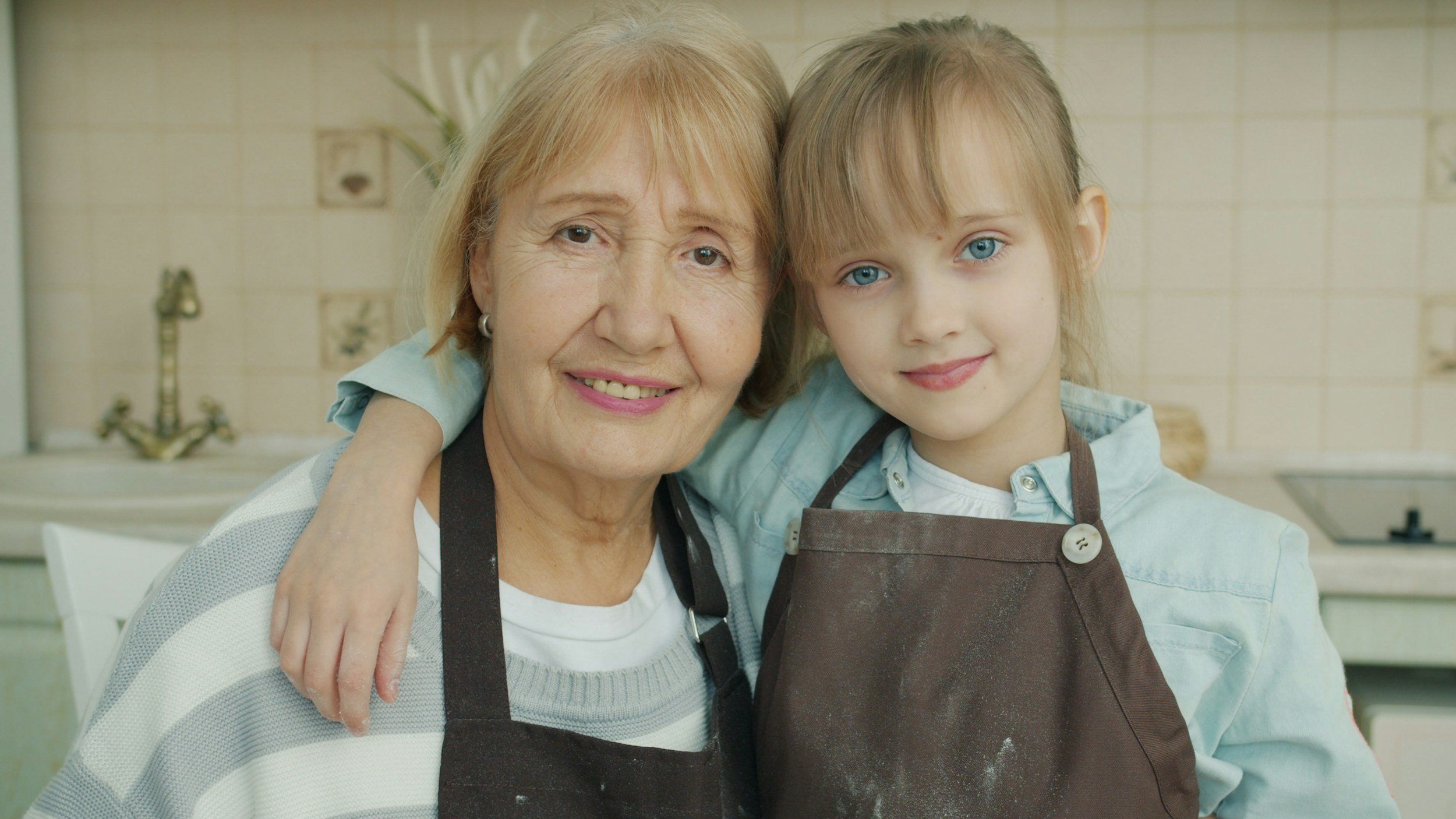 An elderly woman and a young girl with blue eyes hugging and smiling in a kitchen.
