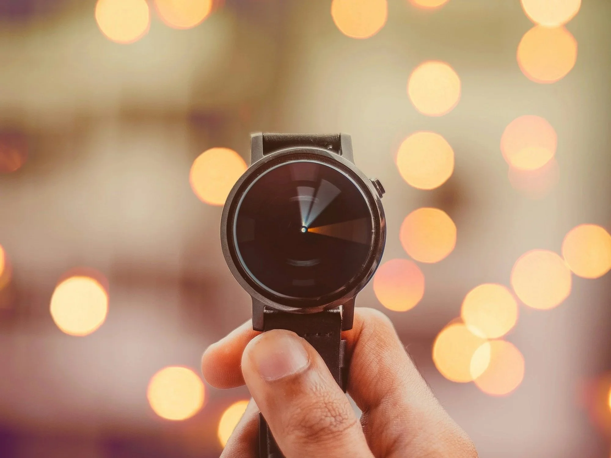 A hand holding a black smartwatch with a round face and a black strap against a blurred background of circular yellow-orange lights.