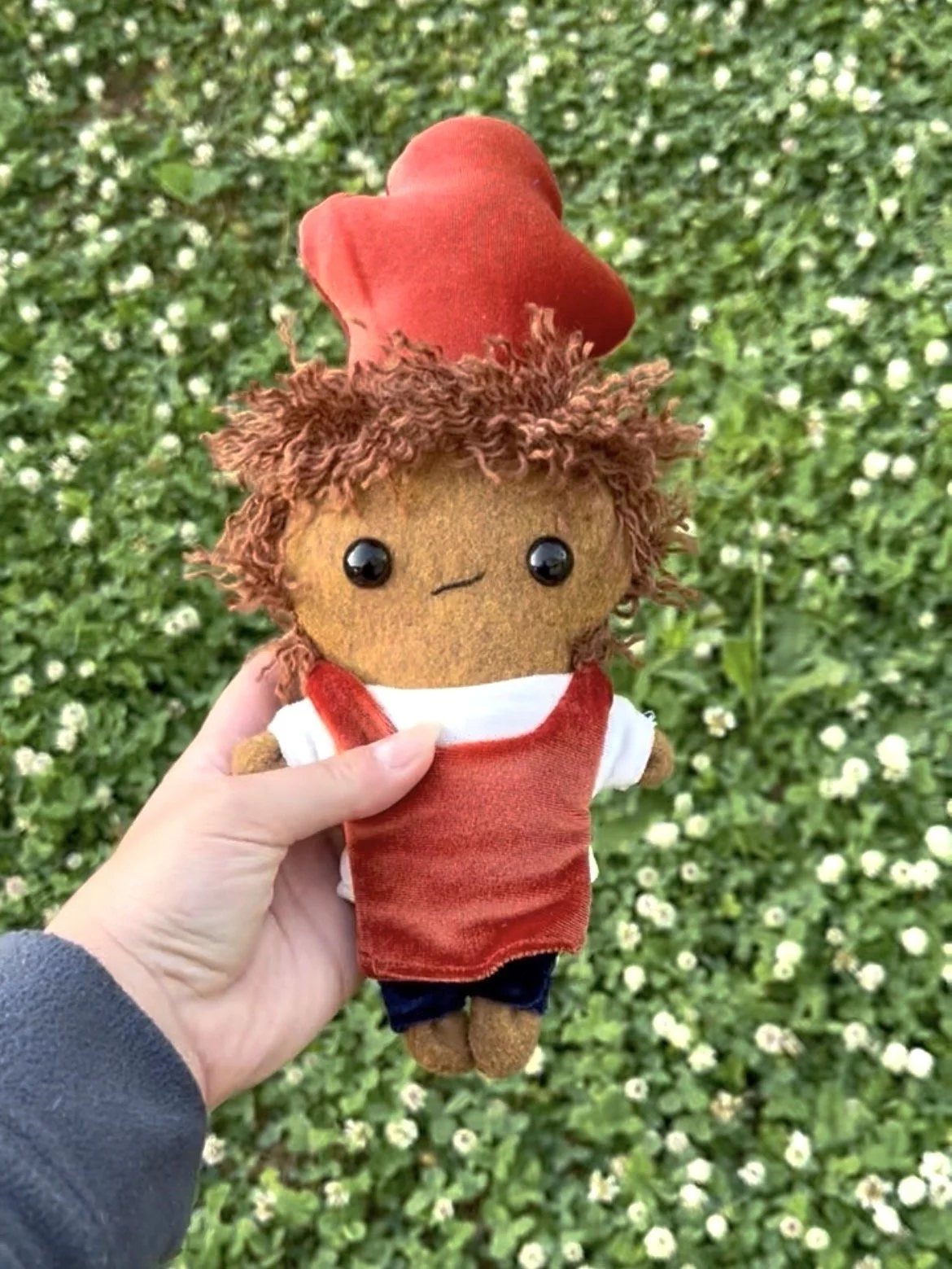 Hand holding a custom handmade plush doll with brown curly hair, black eyes, wearing a red chefs hat, red apron, jeans and white shirt, against a background of green grass.