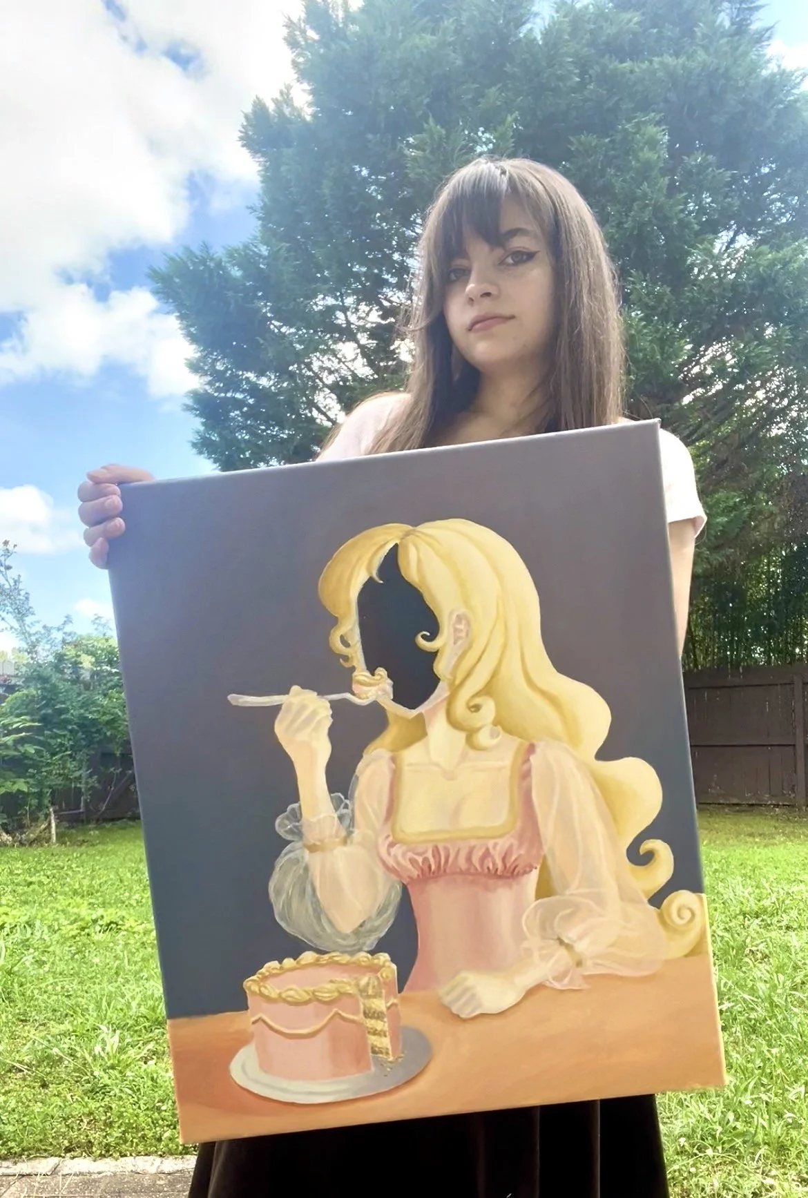 A woman with shoulder-length brown hair holding a soft and surrealist style painting of a girl with long blonde hair, wearing a pink dress, eating cake from a fork, against a background of trees, blue sky, and clouds.