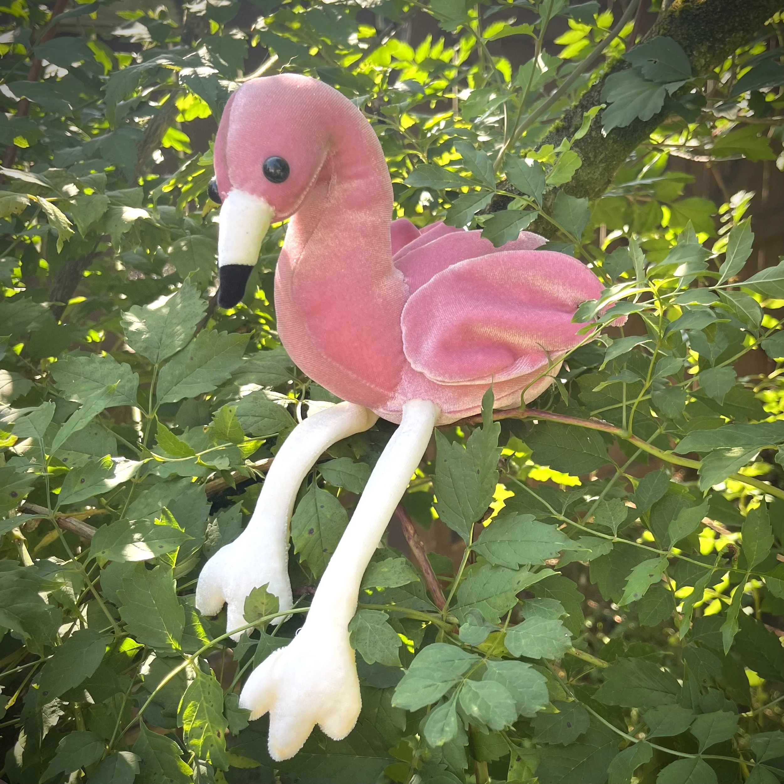 A custom handmade stuffed animal pink flamingo bird toy sitting on a tree branch outside surrounded by green leaves.