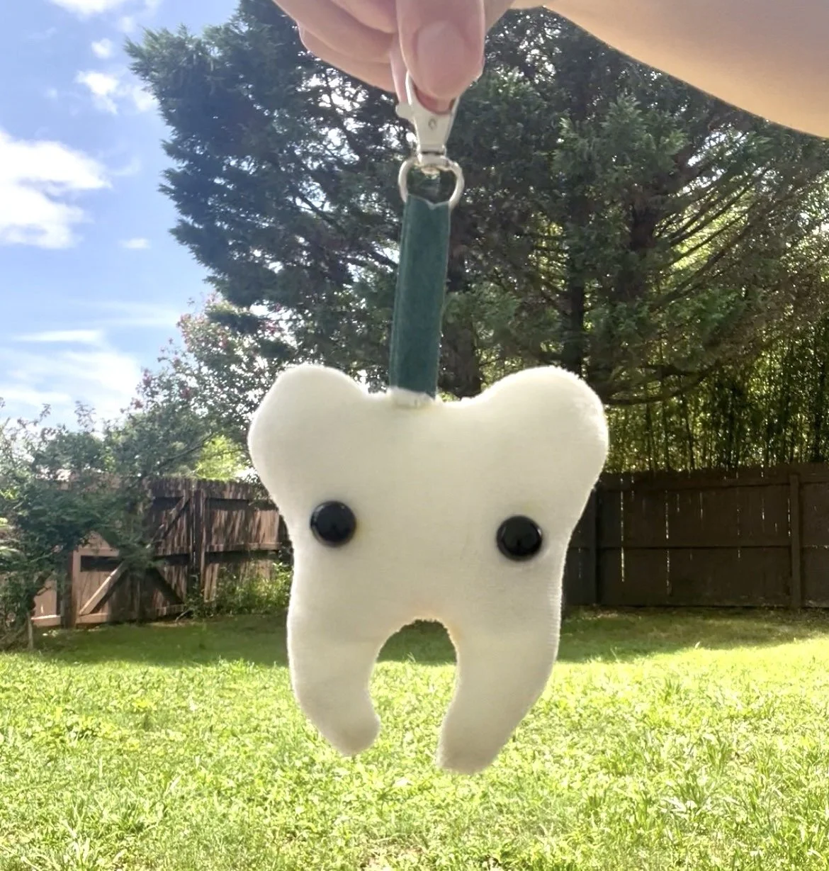 A custom handmade plush toy keychain shaped like a tooth with black eyes and a green ribbon being held outside, with green grass, trees, and a brown fence in the background.