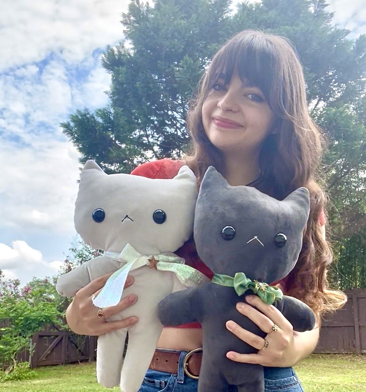 A woman outdoors holding two plush cat dolls, light grey-colored with a ribbon and star, and one dark gray with a green bow, with trees and a fence in the background and a partly cloudy sky.