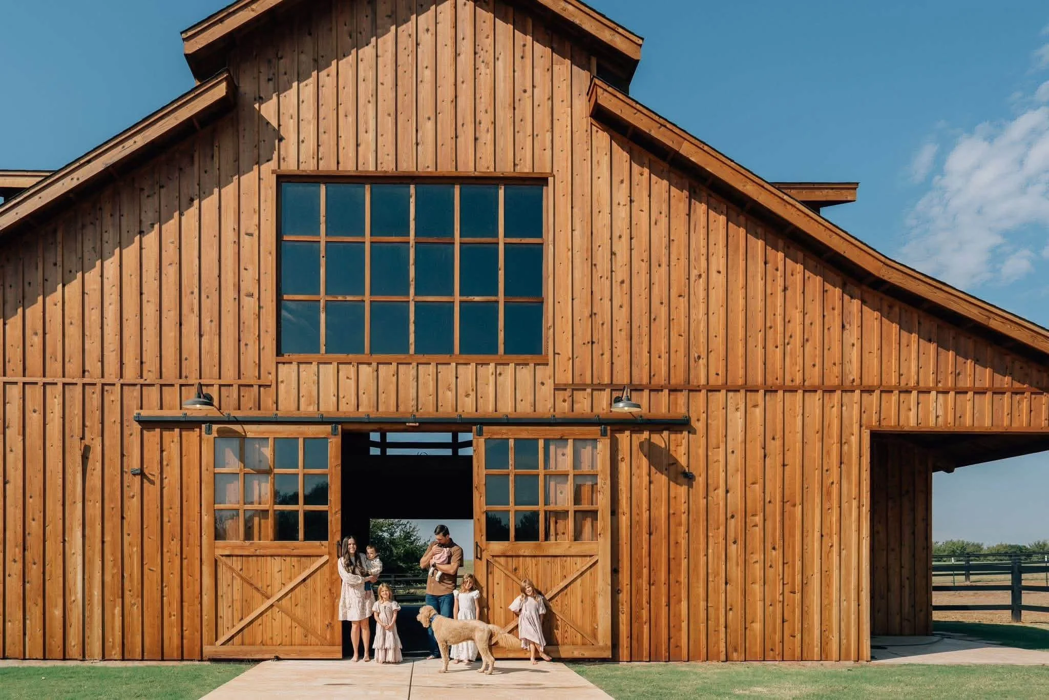 A large wooden barn with sliding doors and a big window on the upper level, under a clear blue sky. Several people and children, along with a dog, are standing near the barn entrance.