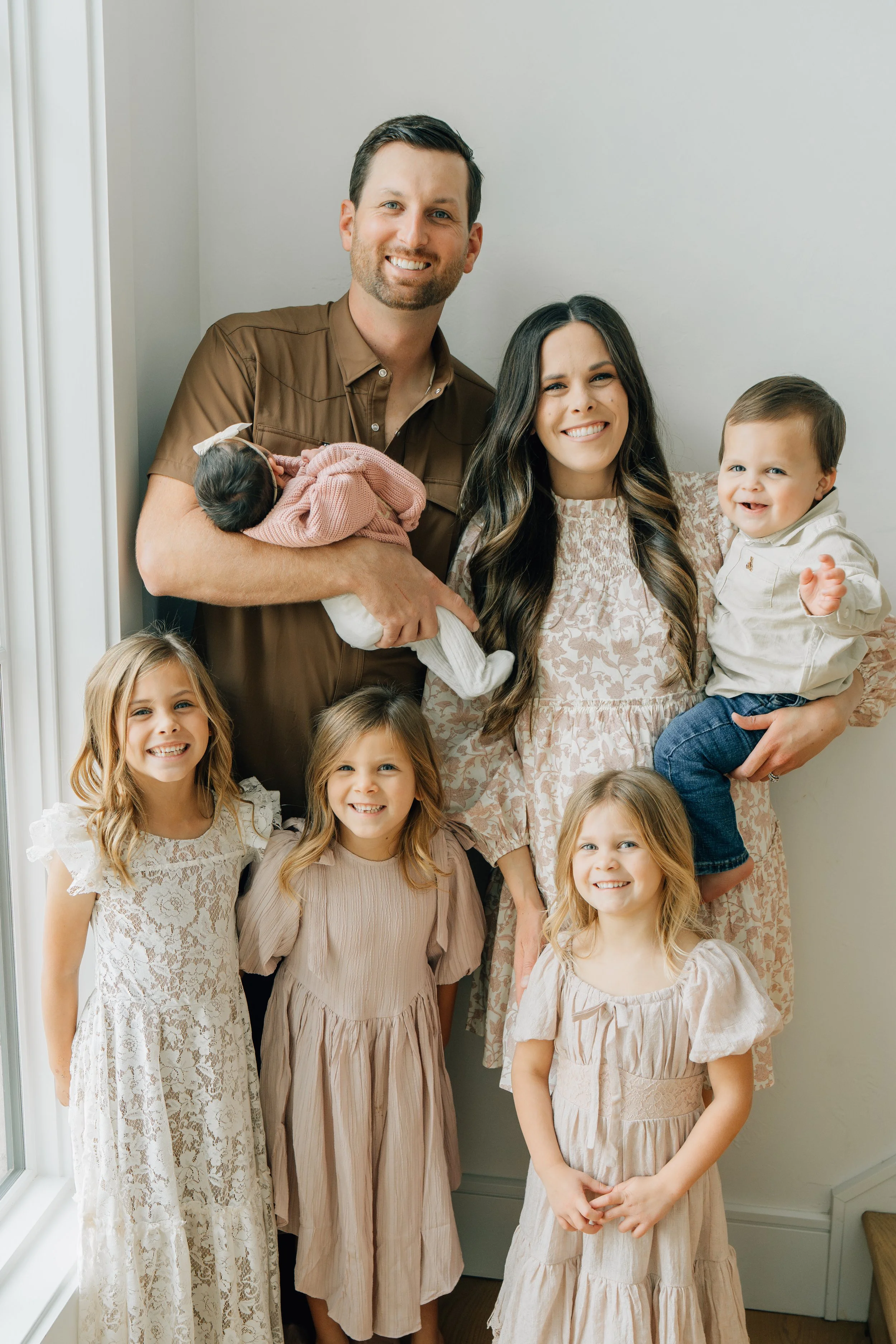 A family of nine posing indoors near a window. The adult man is holding a newborn baby, while the woman and children smile at the camera.