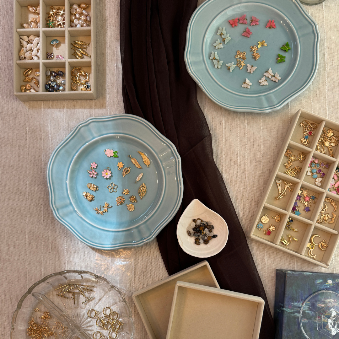 Jewelry display with various charms, pins, and accessories arranged on a table, including plates with small decorative items and organized compartment boxes containing jewelry. Blue and brown table display. Vintage Coastal Cowgirl theme.