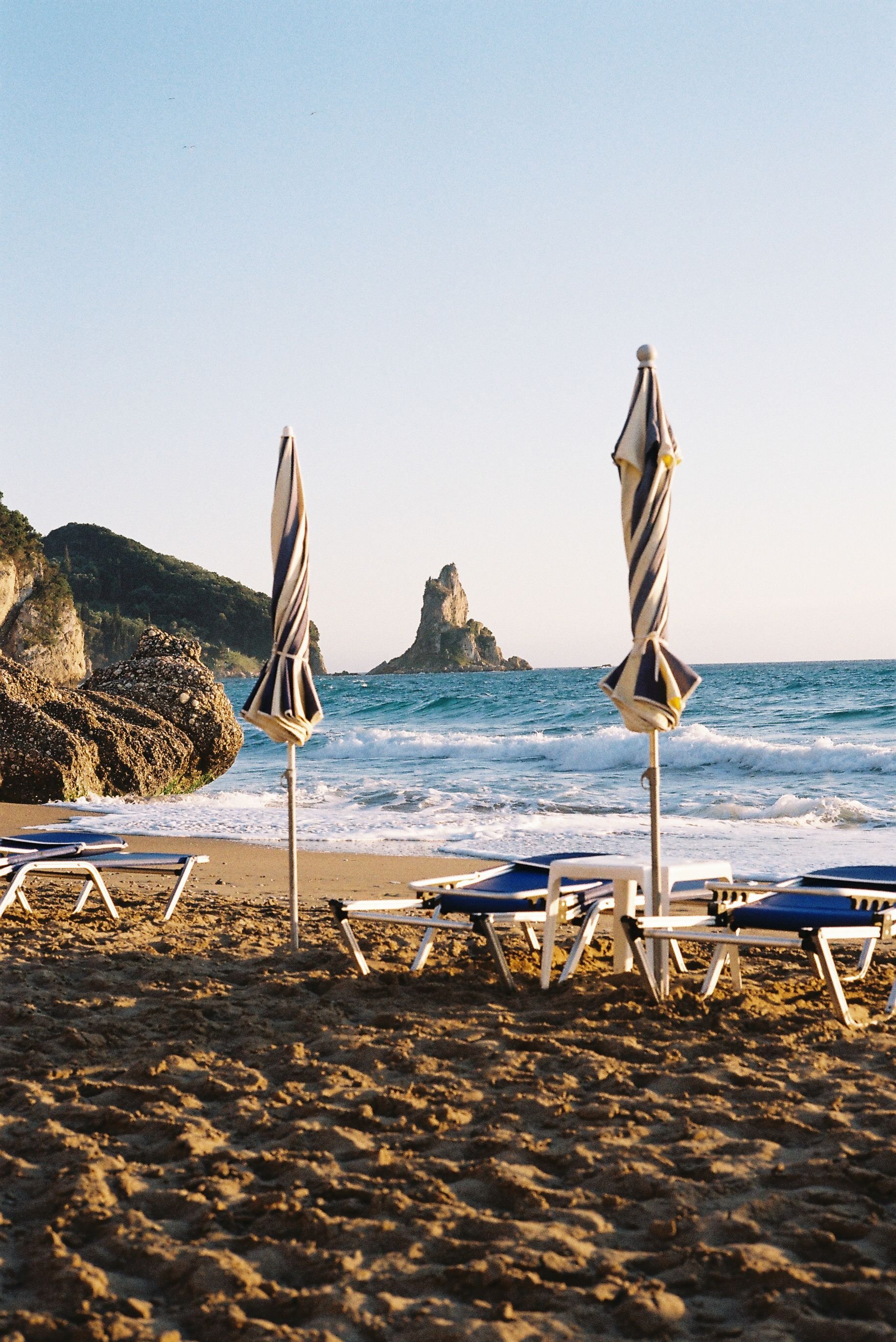 Empty beach with lounge chairs and striped umbrellas facing the ocean, rocky shoreline on the left, large rock formation in the water in the distance, clear sky.
