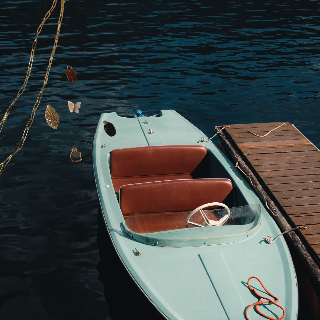 A vintage small boat tied to a wooden dock on calm water, with decorative metallic charms hanging from a chain on the left side, creating a charming, coastal, luxury mood board.