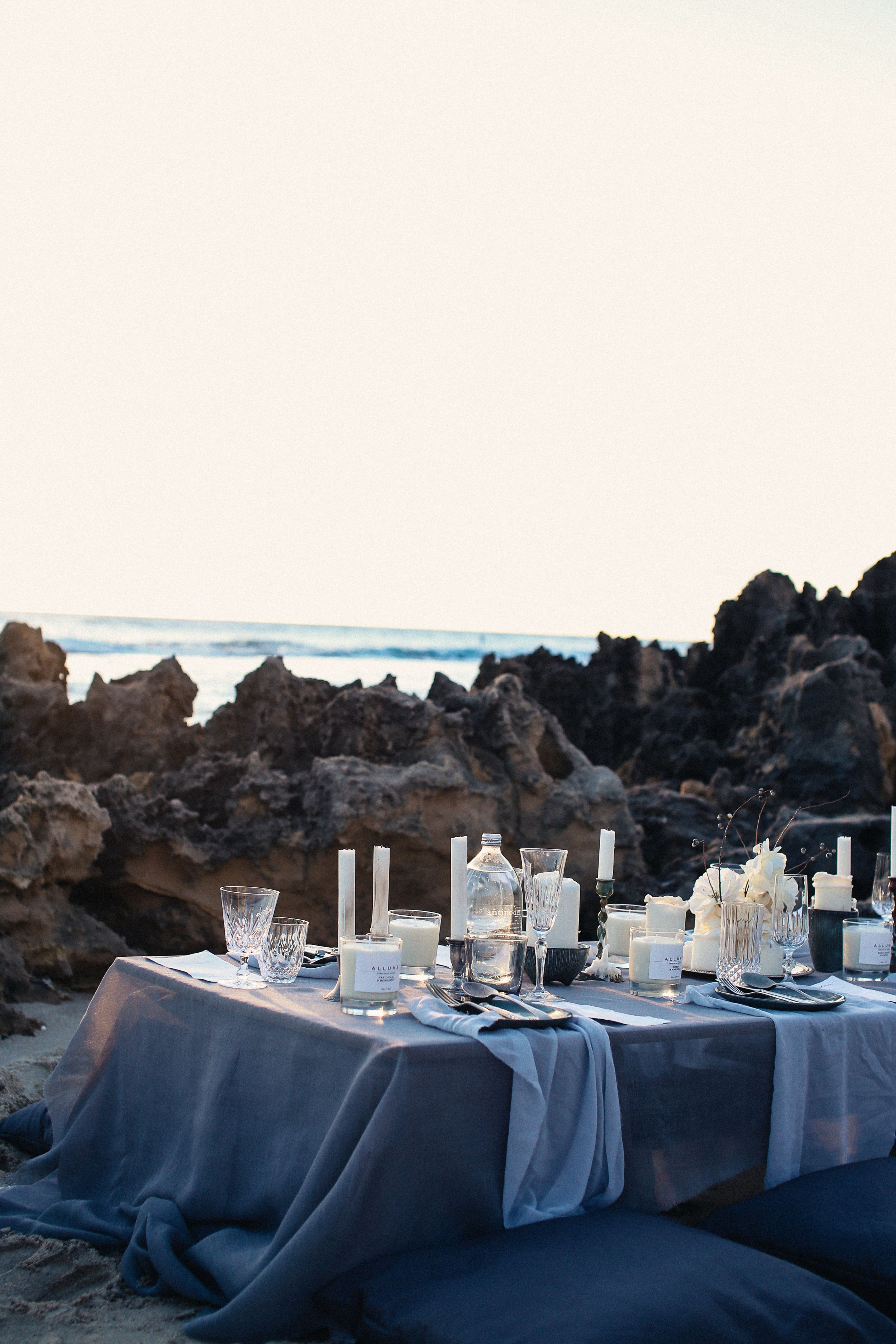 Table set with white candles, glassware, and white flowers on a tablecloth, located near a rocky beach with ocean waves in the background during sunset or dusk.