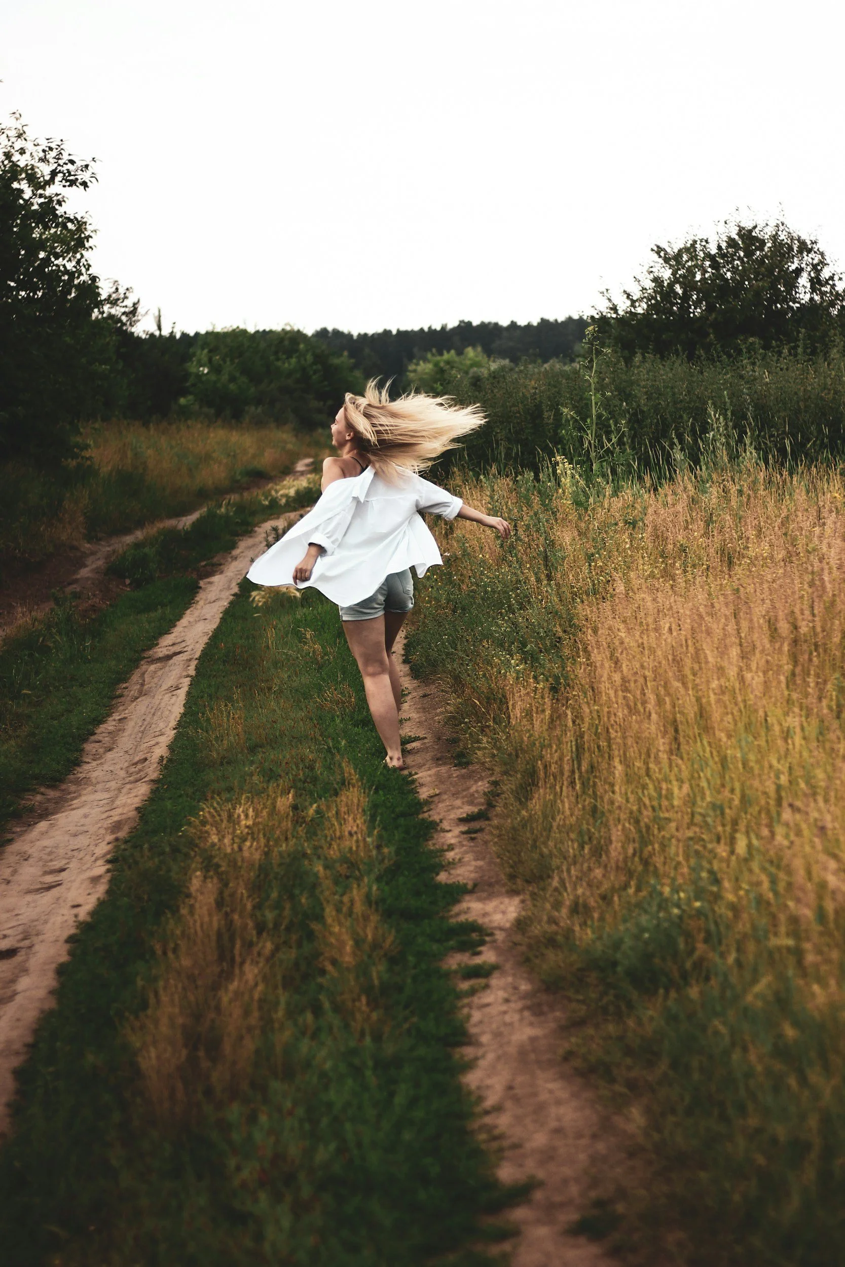 A woman with long blonde hair wearing a white shirt and shorts is running on a dirt path through a field of tall grass and wildflowers in a rural landscape.