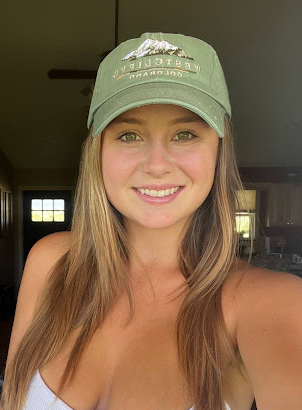 Young woman taking a selfie indoors, wearing a green baseball cap and a white tank top, smiling.