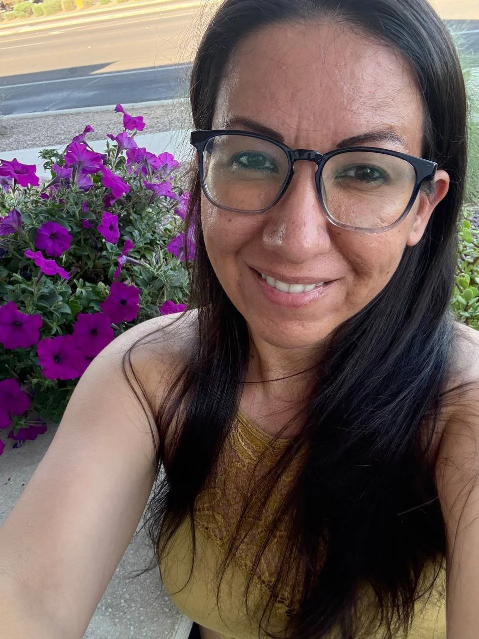 A woman with long dark hair and glasses smiling for a selfie outdoors near purple petunias.