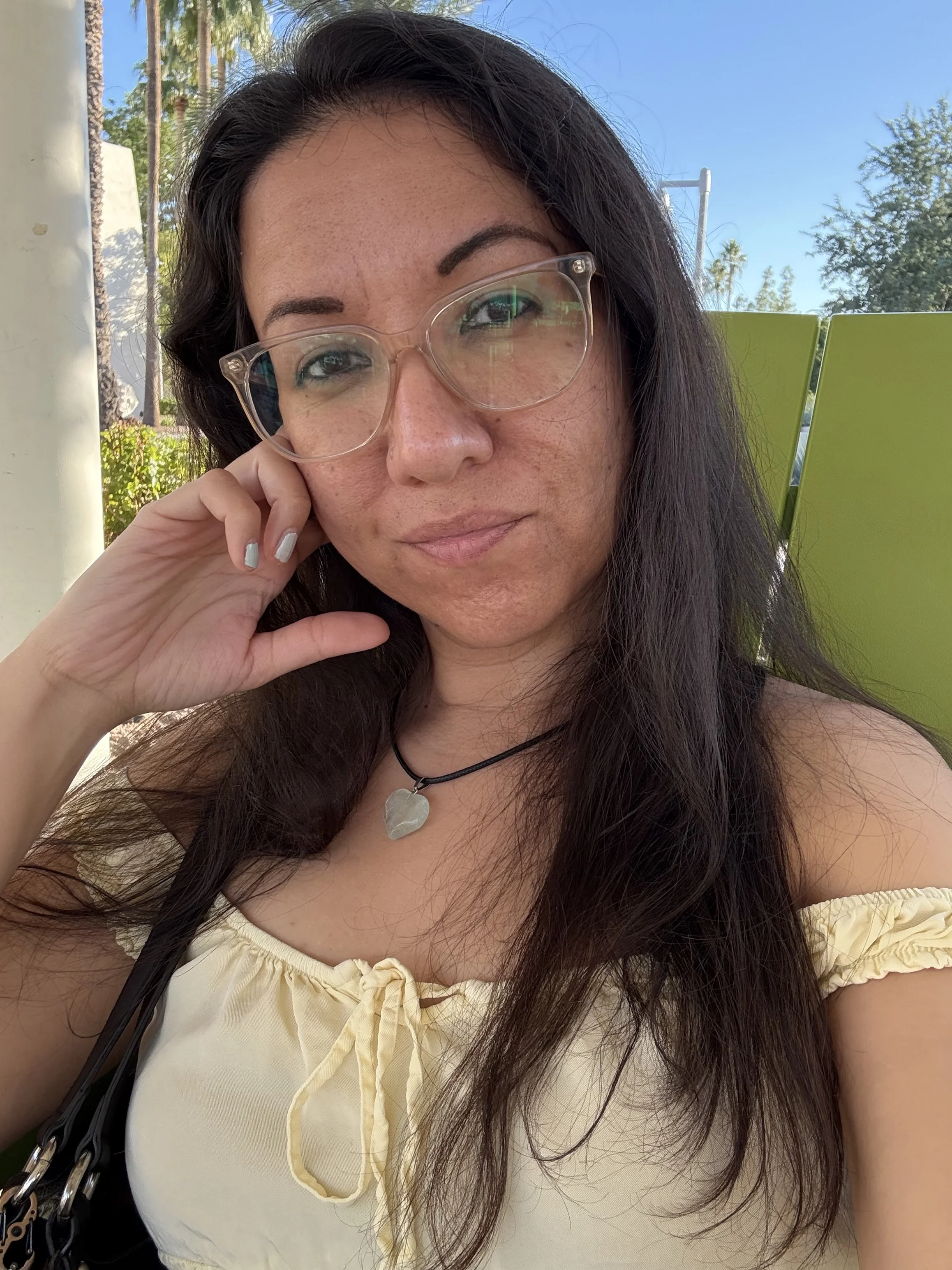 Close-up of a woman with long dark hair and glasses, smiling gently at the camera, sitting outdoors in bright sunlight, with trees and a blue sky in the background.