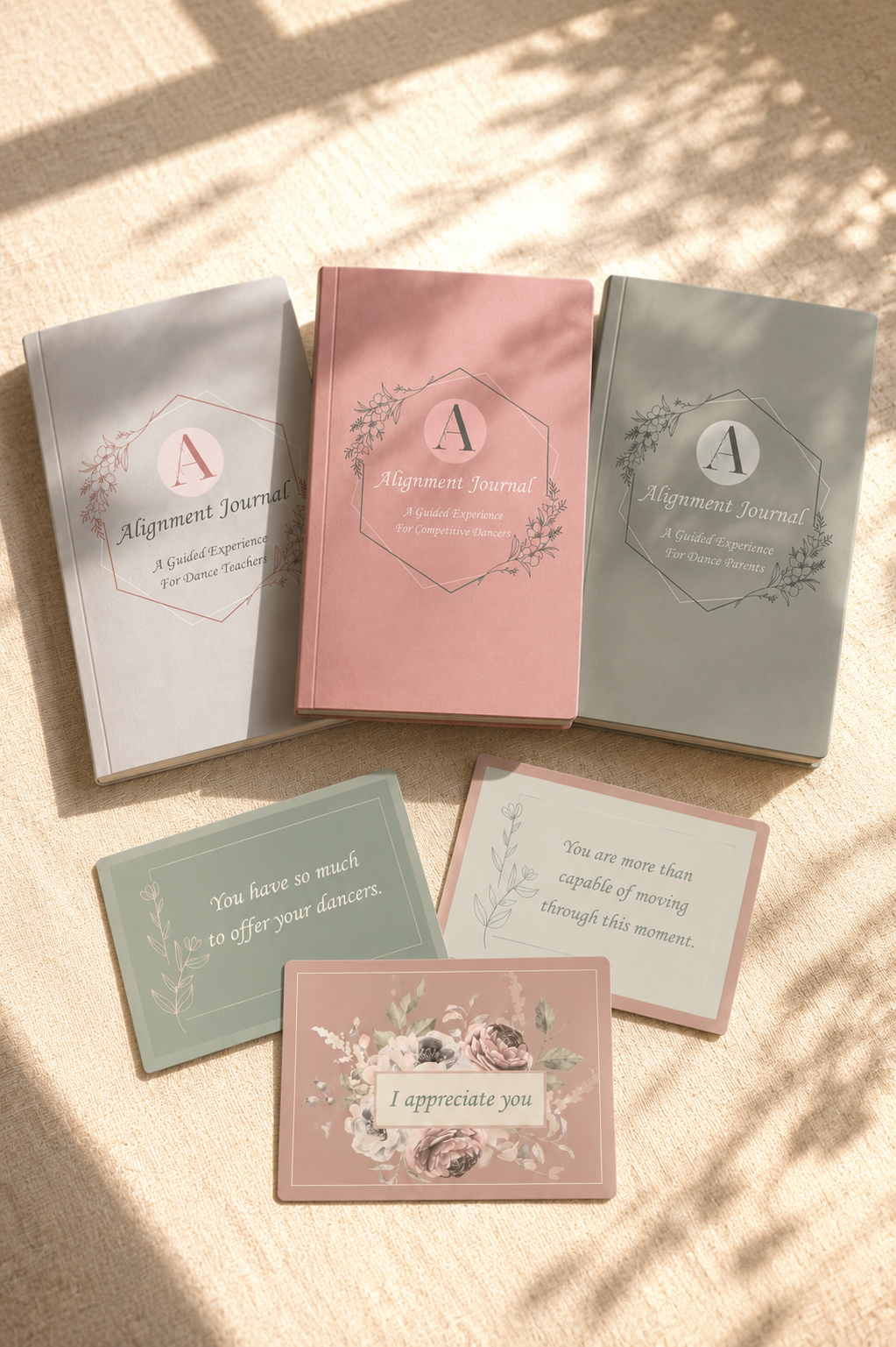 Three softcover books titled 'Alignment Journal' for dance teachers, dance students, and dance parents, with pastel-colored covers in white, pink, and gray. In front, three cards with motivational messages and floral designs are displayed on a beige fabric surface illuminated by sunlight.