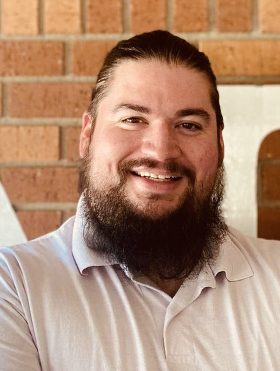 A smiling man with a beard and slicked-back hair, wearing a light-colored collared shirt, standing in front of a brick wall.