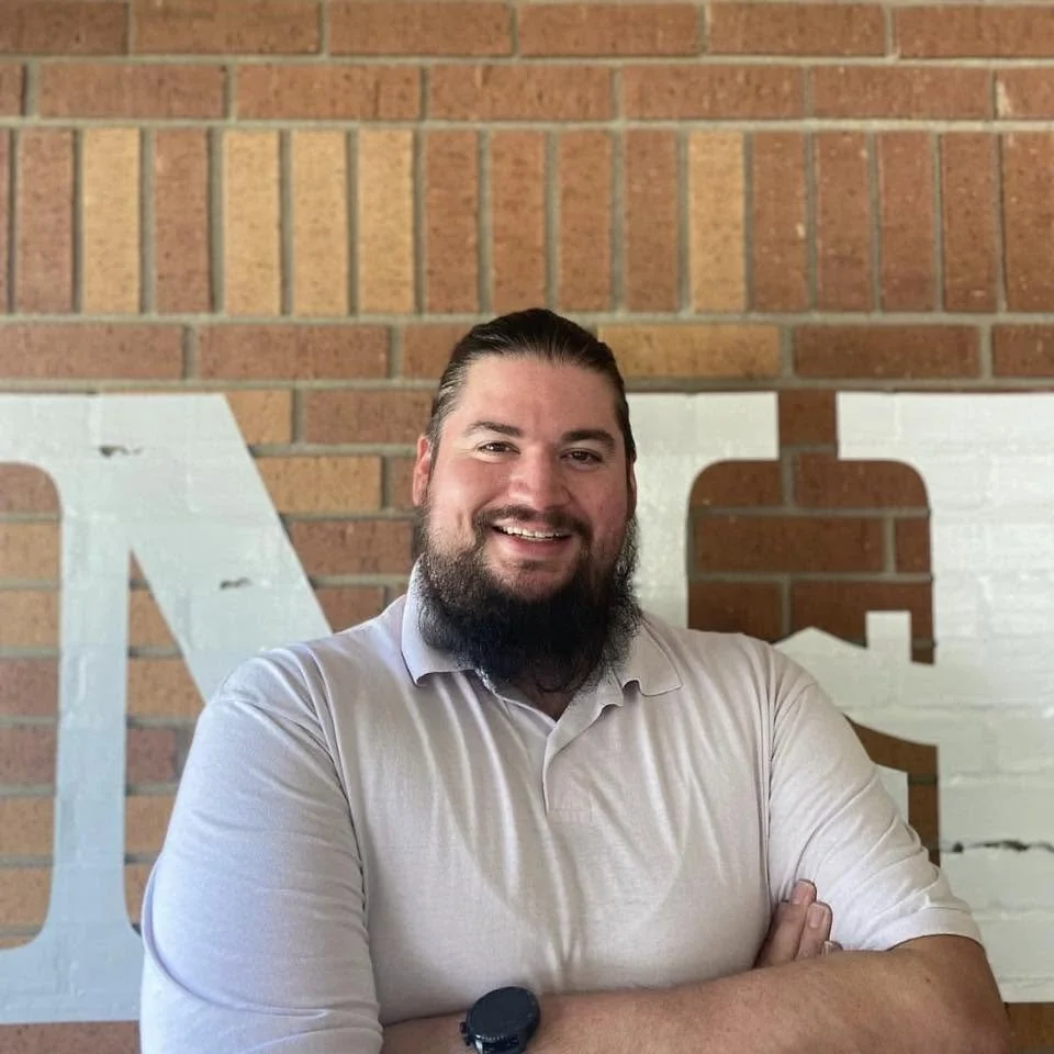 Man with dark hair and beard smiling, standing in front of a brick wall with large white letters partially visible.