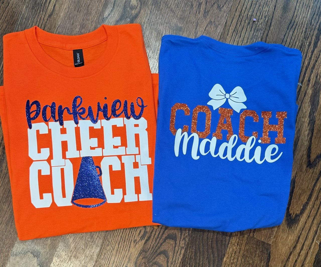 Two folded T-shirts on a wooden floor, one orange with the text 'parkview CHEER CUCH' and a blue party hat graphic, the other blue with the text 'COACH Maddie' and a white bow graphic.