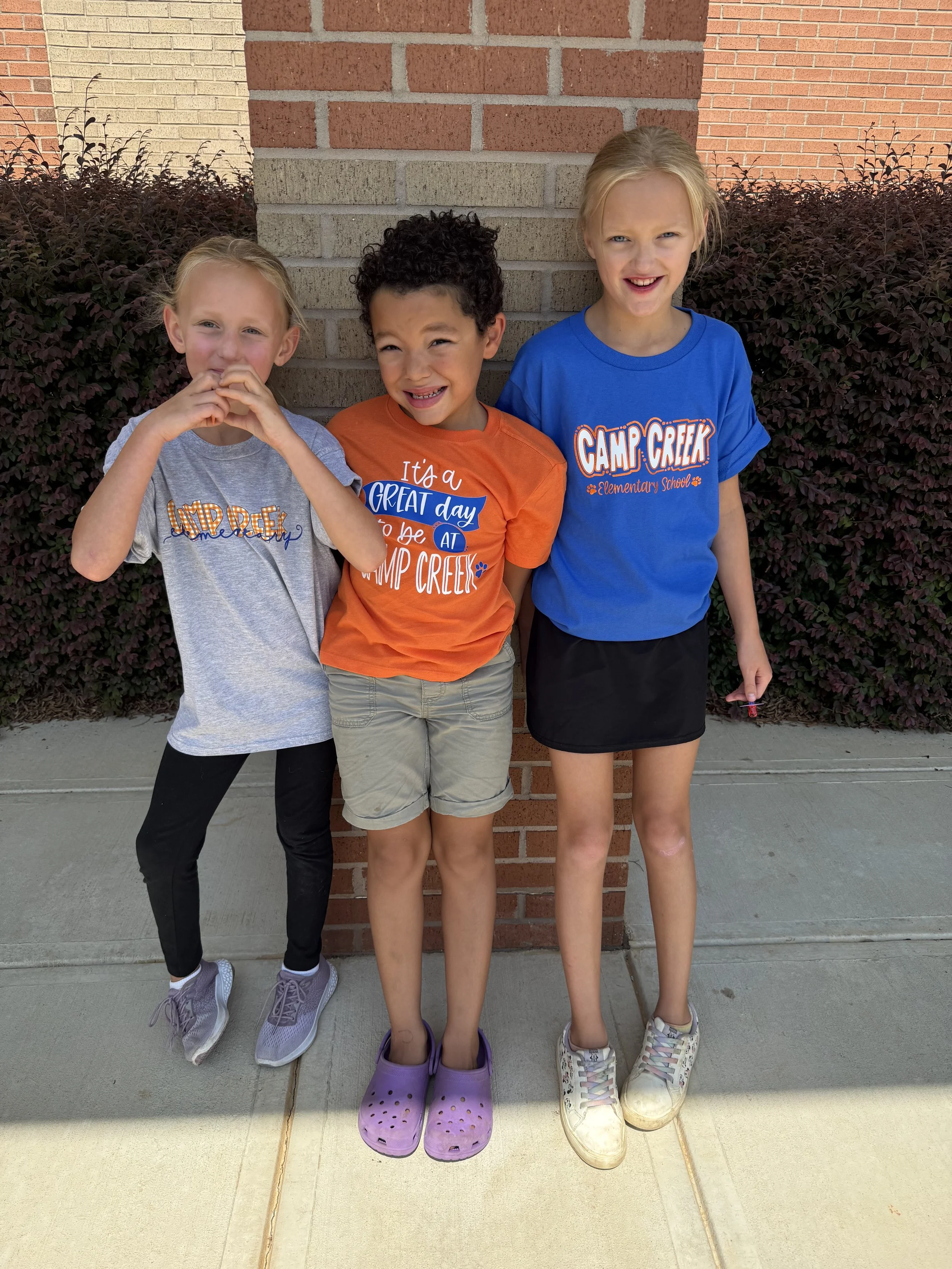 Three children standing outside in front of a brick wall with purple and white shoes and casual clothing, smiling and posing for the photo.
