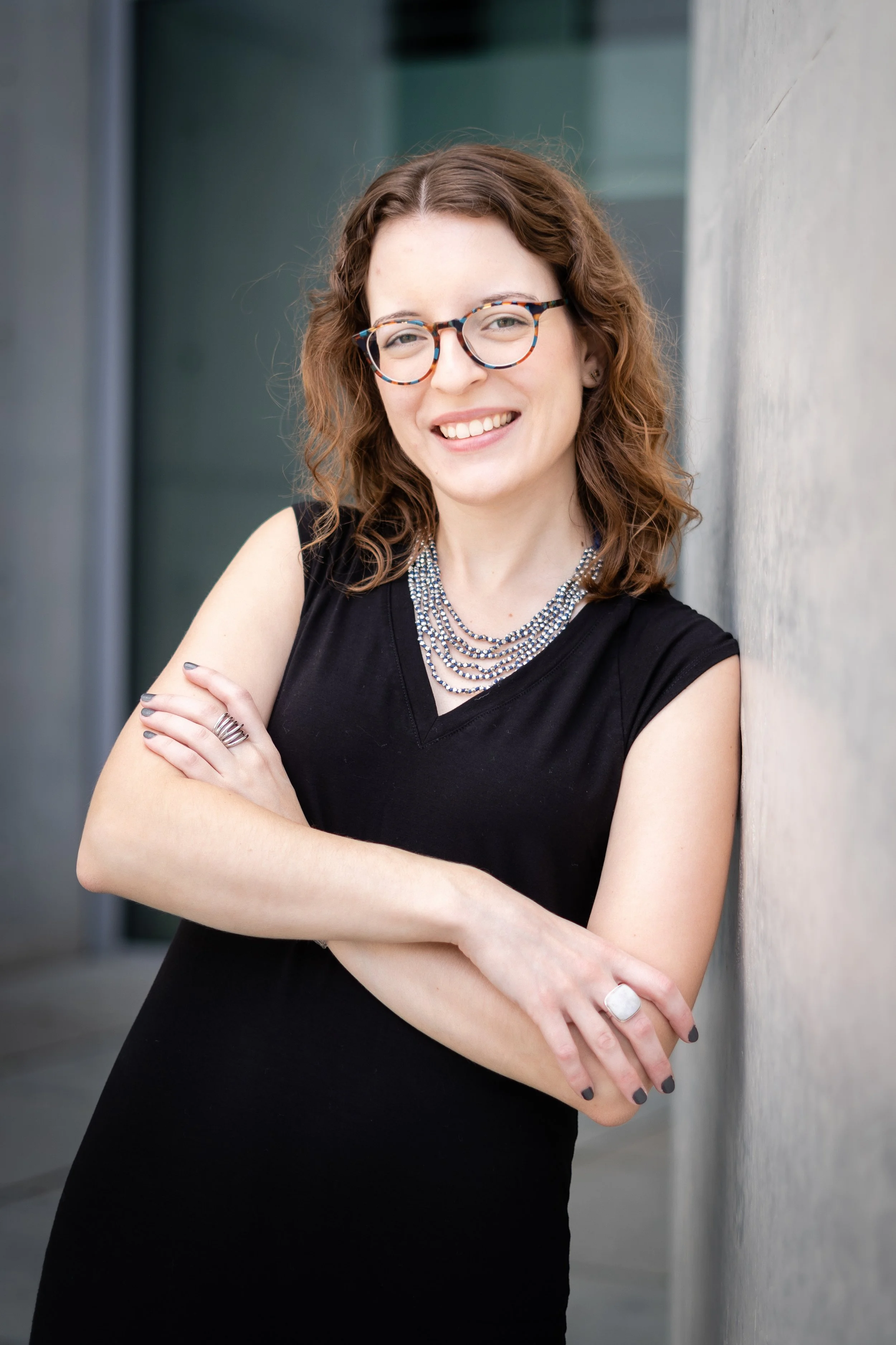 A photograph of Holly Yanacek in Berlin. She has curly brown hair and is wearing glasses, a black dress, layered necklace, and rings, smiling and leaning against a wall outside.