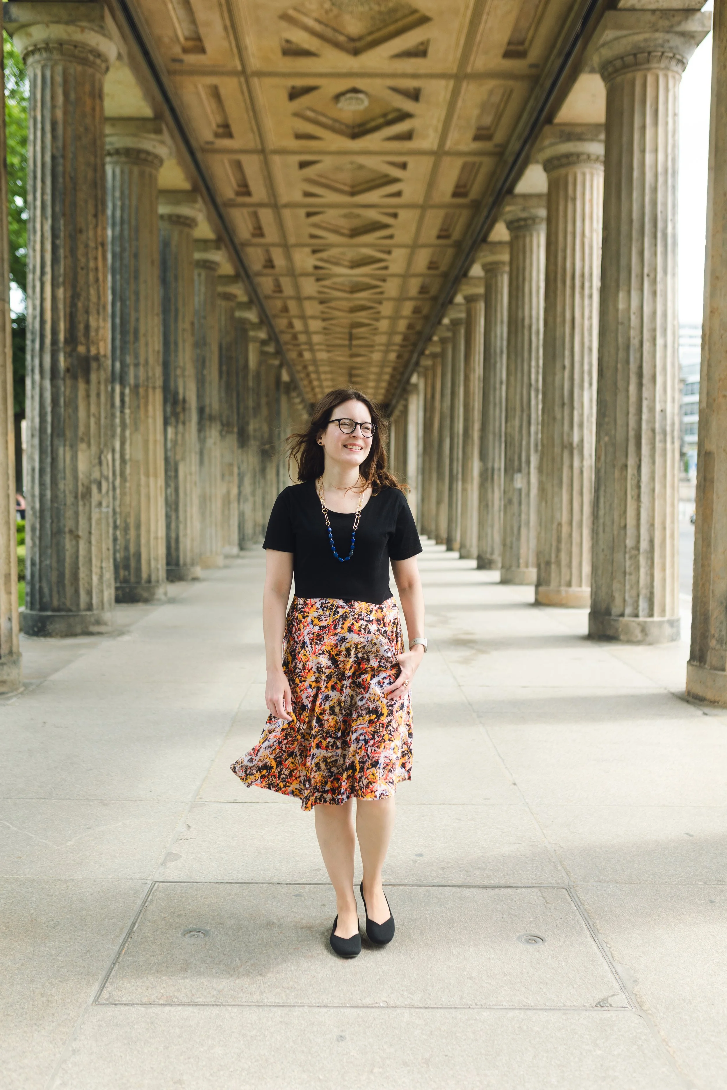 A woman walking under a colonnade with columns on either side, wearing a black top, a colorful patterned skirt, black shoes, glasses, and a blue beaded necklace, smiling.