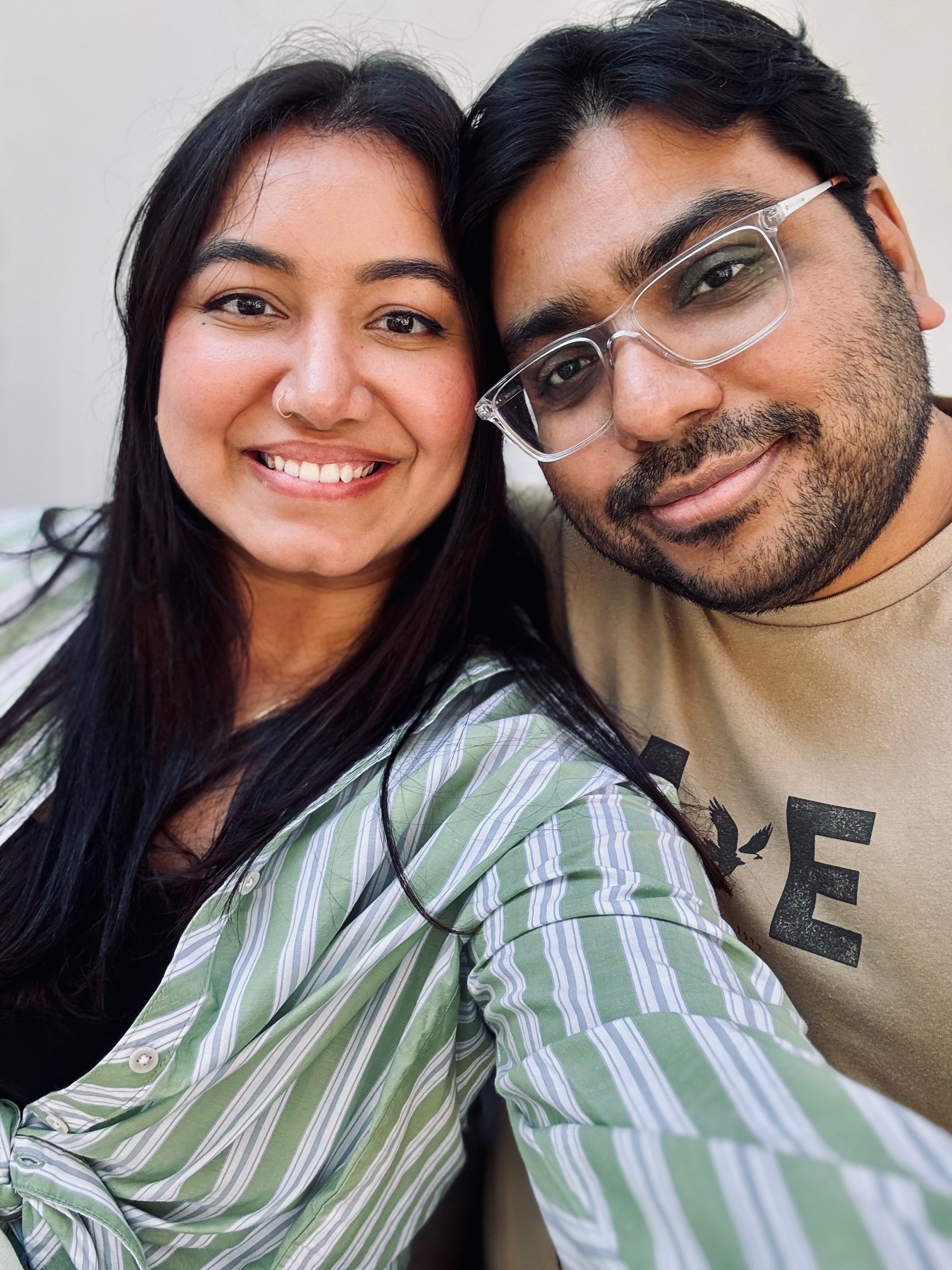 Two smiling people taking a close-up selfie, a woman with long dark hair and a man with dark hair and glasses.