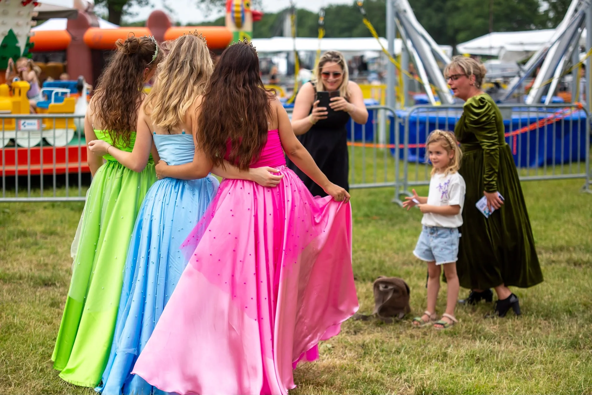 Vrouwen in kleurrijke avondjurken die elkaar omarmen op een kermis, met een kleine meisje en een vrouw die op de achtergrond staan. Er zijn attracties en een hek te zien op de achtergrond.