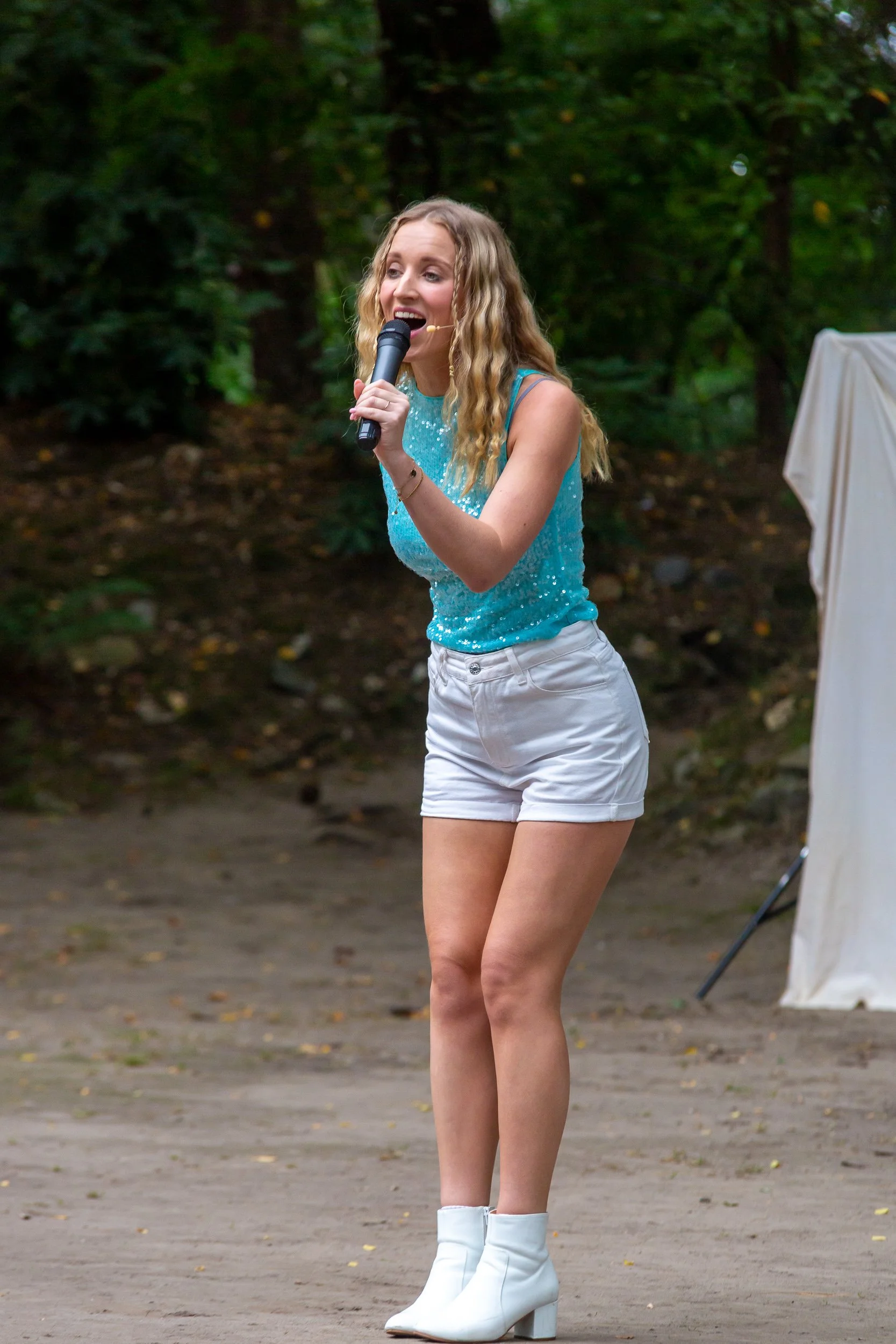 Een jonge vrouw met lang, golvend blond haar draagt een glanzende blauwe top en witte korte broek. Ze staat buiten op een pad met een groene bos op de achtergrond, houdt een microfoon vast en lijkt te zingen of spreken.