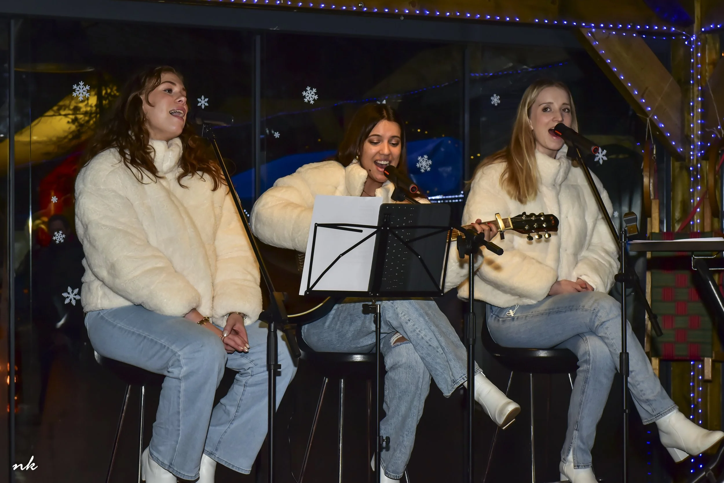 Drie vrouwen zingen en spelen gitaar tijdens een kerstshow met blauwe kerstverlichting en sneeuwvlokken in de achtergrond.